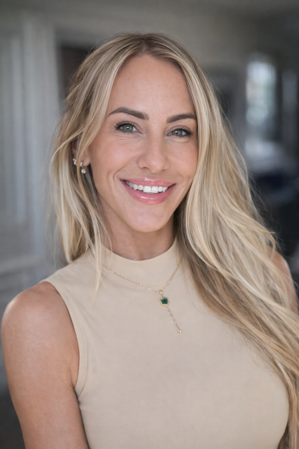 Close-up of a smiling woman with long blonde hair, wearing a beige sleeveless top, jewelry, and natural makeup.