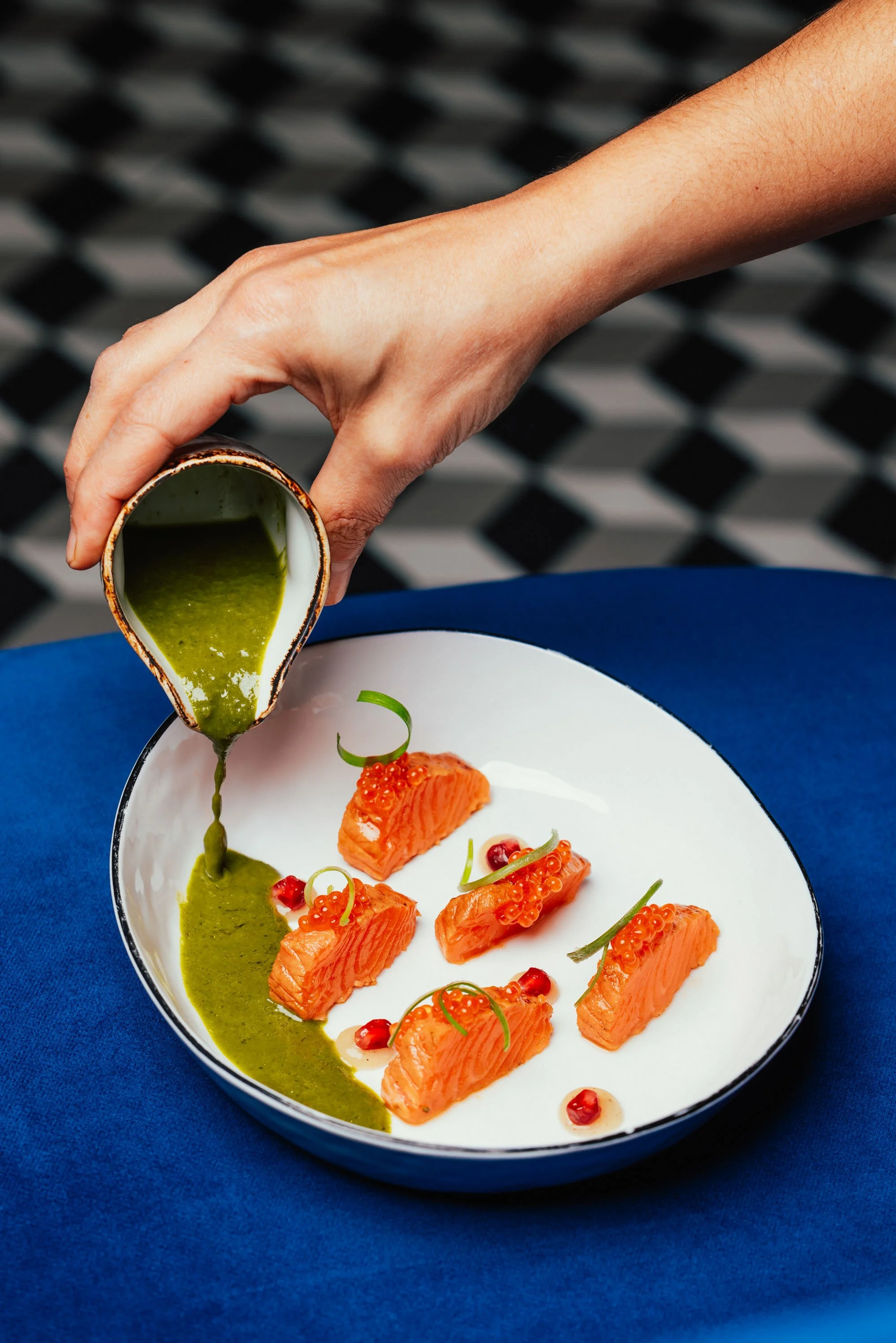 Hand gießt grüne Soße über mehrere Stücke rohen Lachses auf einem weißen Teller, dekoriert mit roten Beeren und grünen Garnierungen, auf einem blauen Tisch.