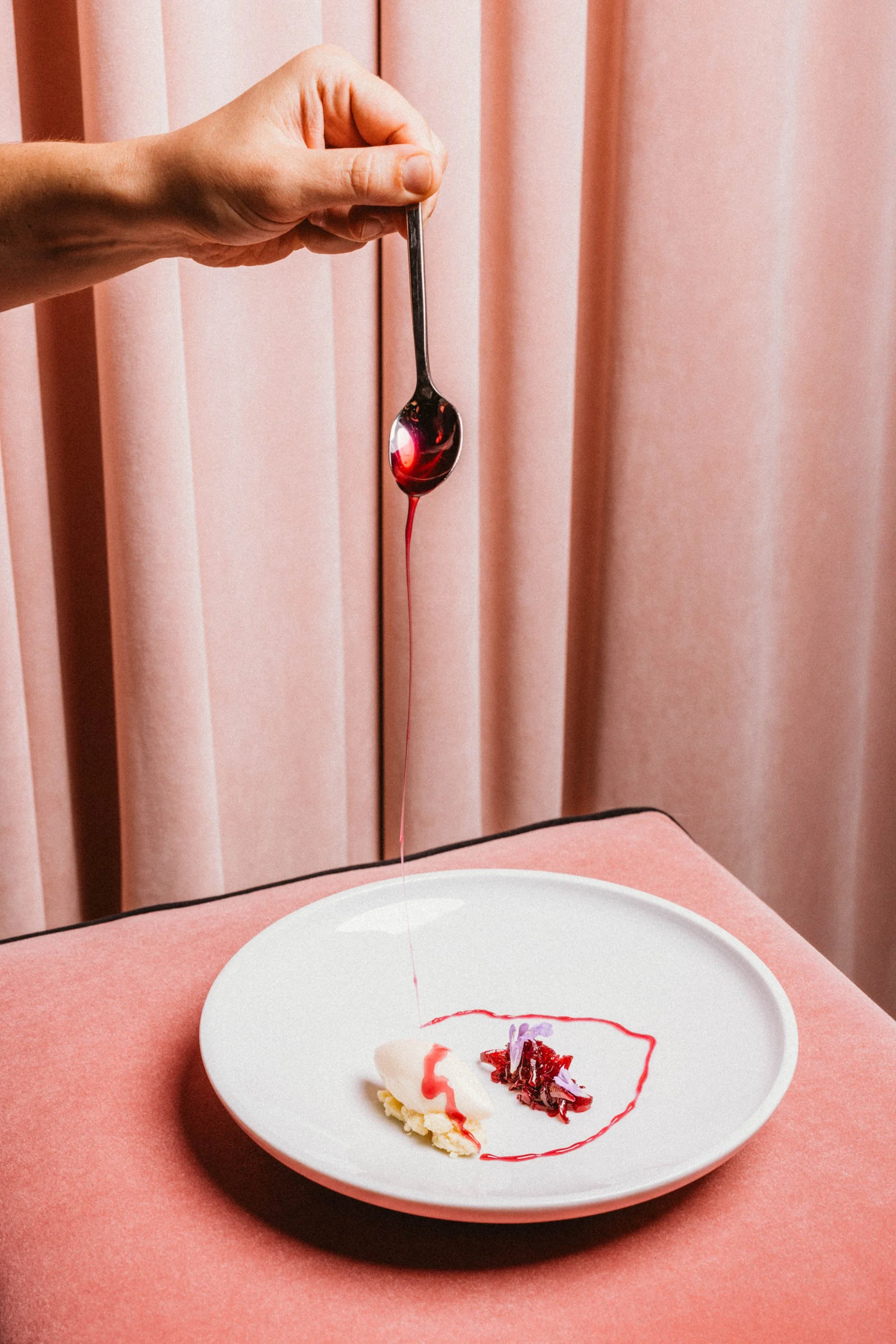 Hand hält einen Löffel mit roter Soße über einem Dessert auf einem weißen Teller, das mit roter Soße garniert ist. Der Hintergrund besteht aus rosa Vorhängen, und der Teller liegt auf einem rosa Tuch.