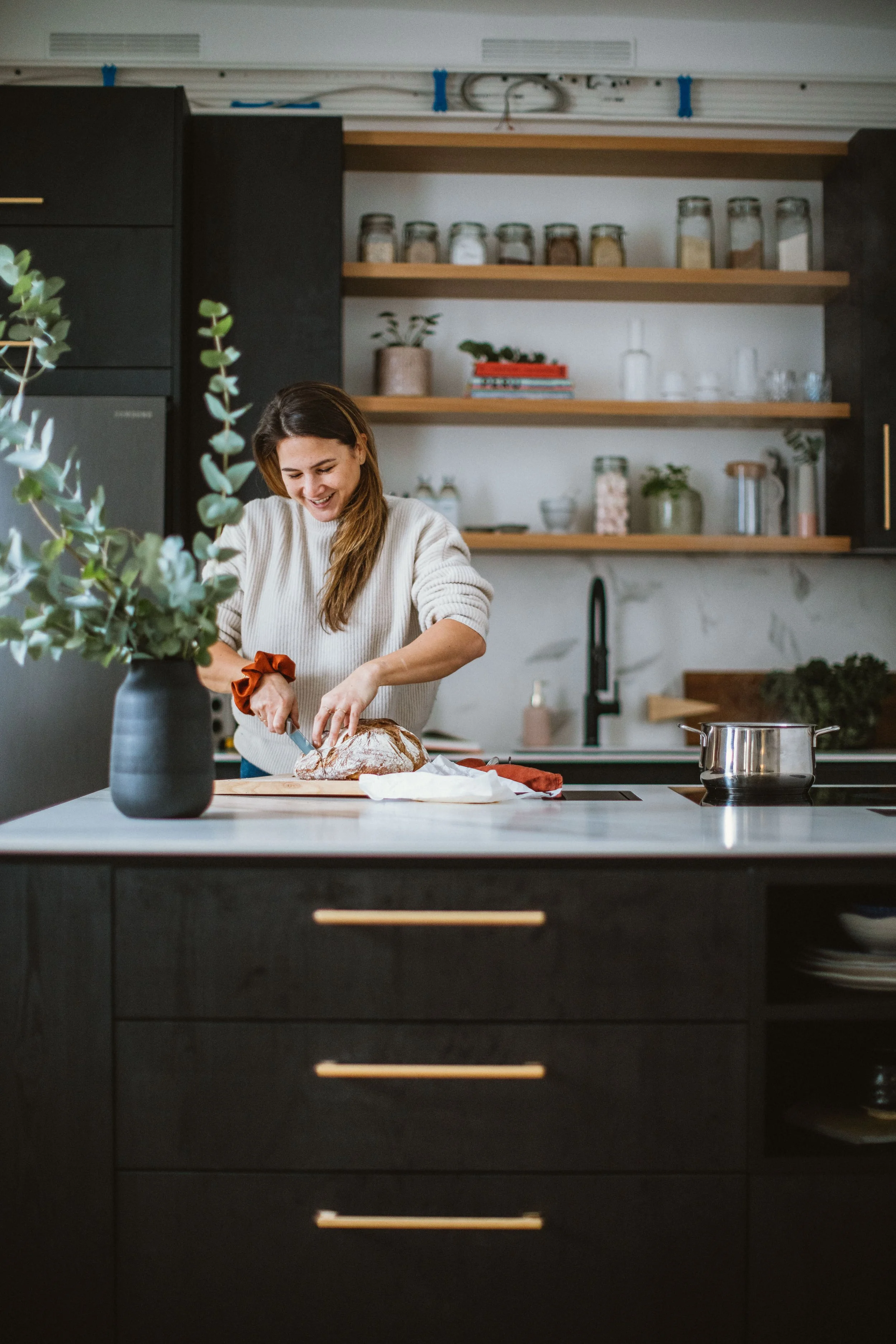 Eine Frau schneidet in einer modernen, schwarzen Küche ein Brot, während sie lächelt. Es gibt eine Vase mit grünen Zweigen auf der Theke und offene Holzregale im Hintergrund mit Gläsern, Büchern und Pflanzen.