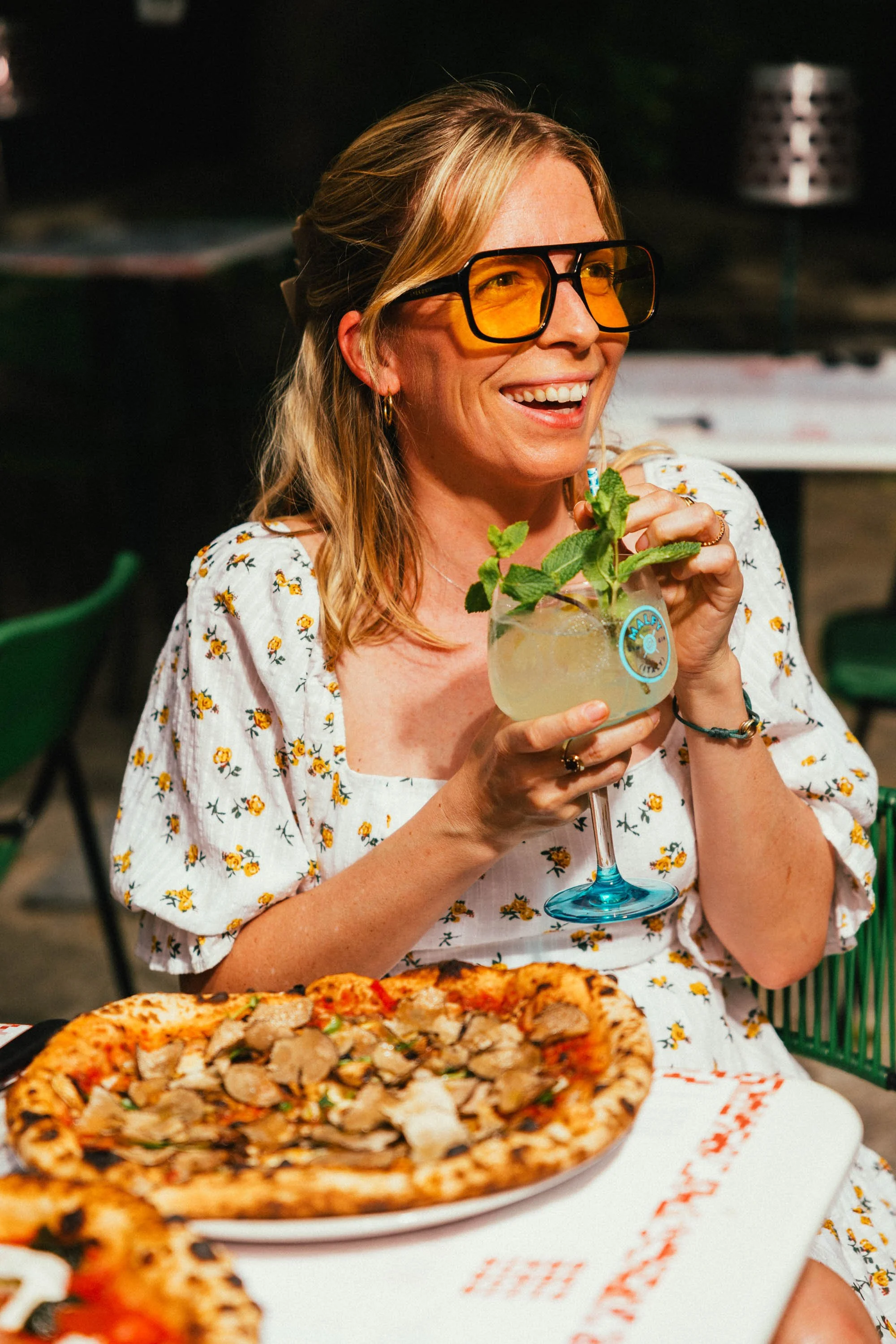 Eine lachende Frau mit blonden Haaren, Sonnenbrille mit gelben Gläsern, trägt ein weißes Kleid mit gelben Blumen, hält ein Getränk mit Minzblättern, sitzt an einem Tisch mit Pizza.
