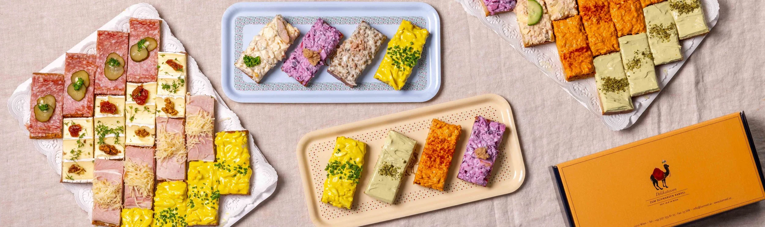 Verschiedene Arten von belegten Broten, darunter Käsebrote, Schinkenbrot, und vegetarische Varianten, schön angerichtet auf Platten und Serviertabletts auf einem Tischtuch.