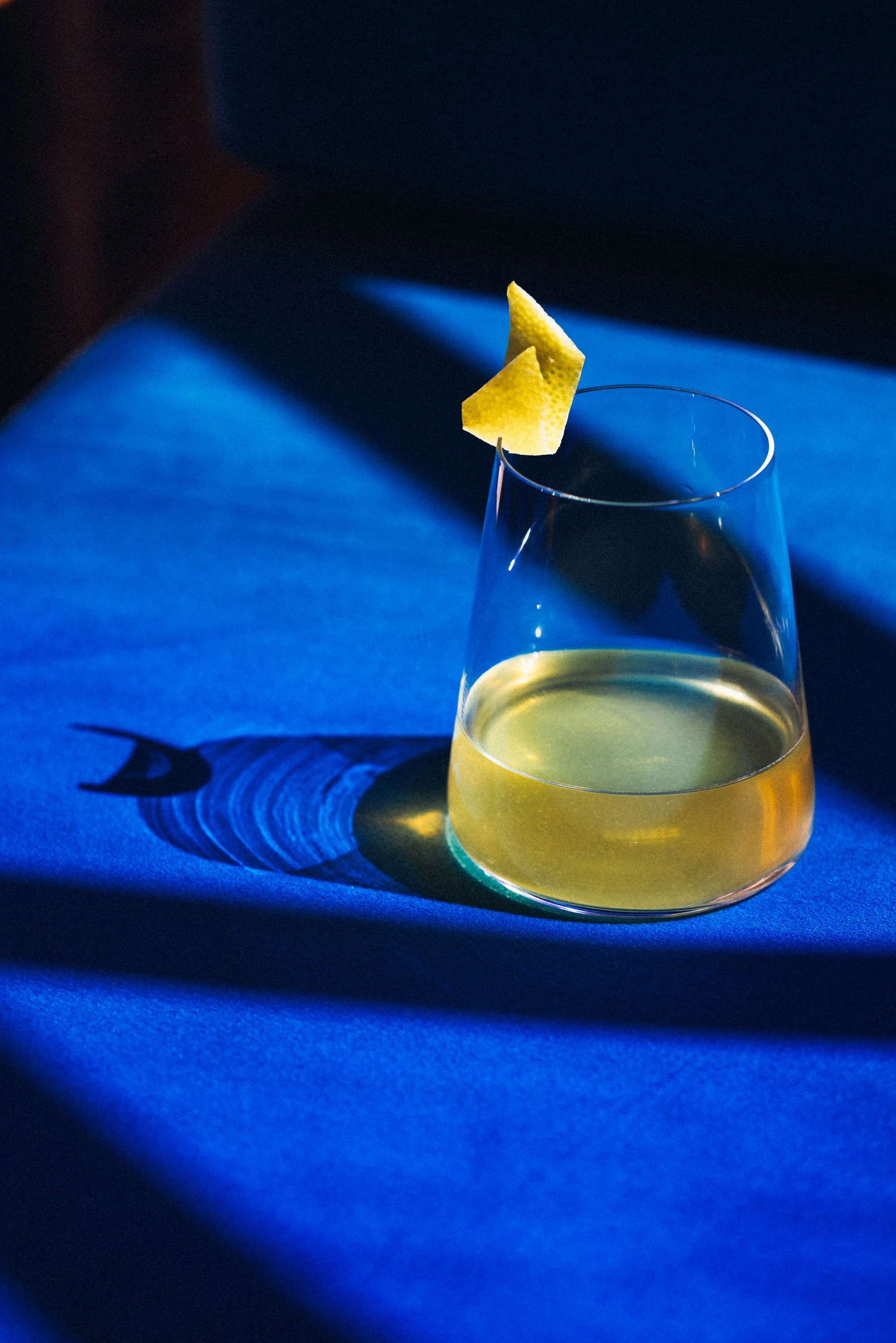 Ein Glas mit gelbem Getränk, garniert mit einer Zitronenscheibe, auf einem blauen Tuch, Schatten auf dem Tuch