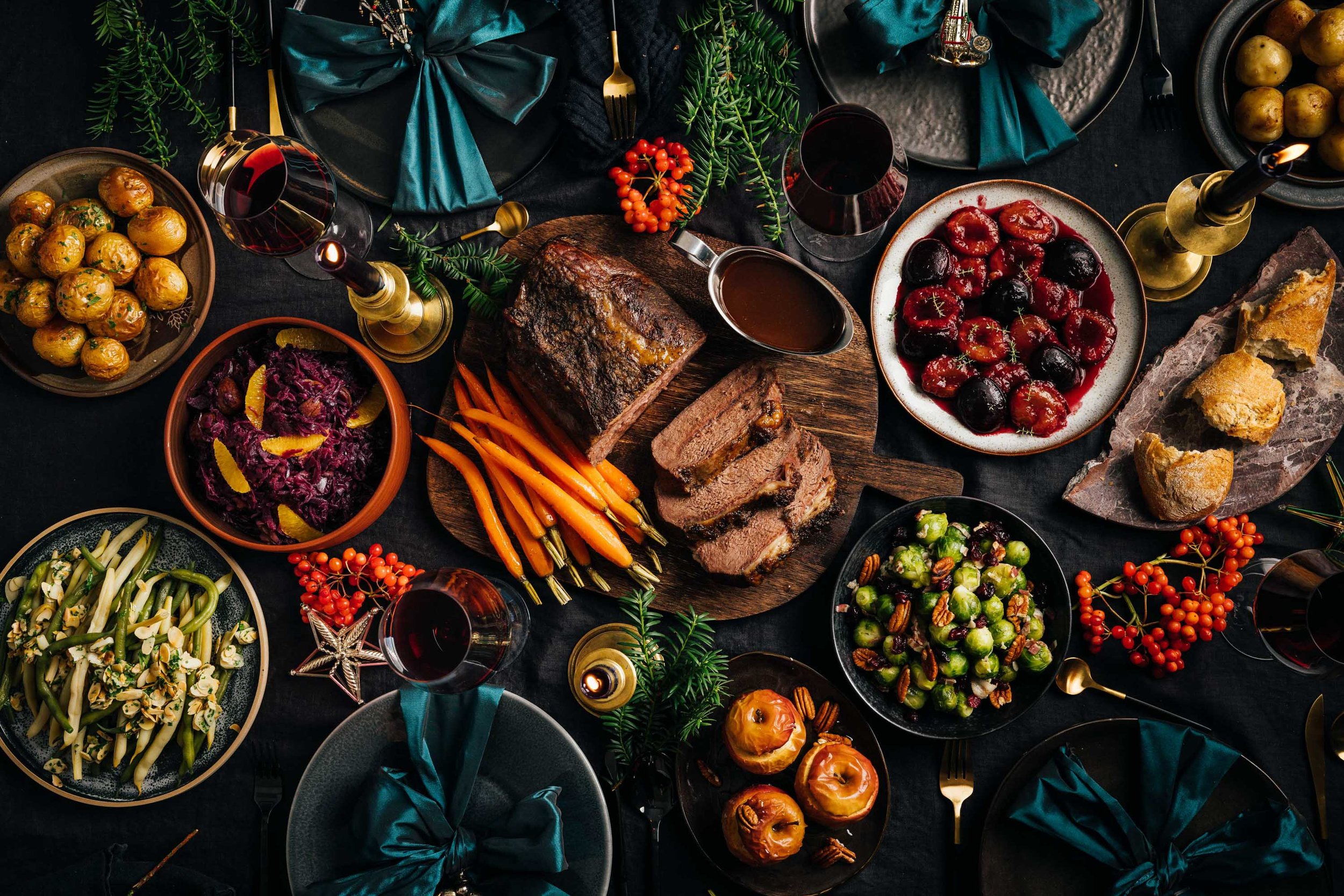 Festlich gedeckter Tisch mit verschiedenen Speisen, including gebratenes Fleisch, Beilagen, Beeren, Gemüse, Wein, Kerzen und Dekorationen.