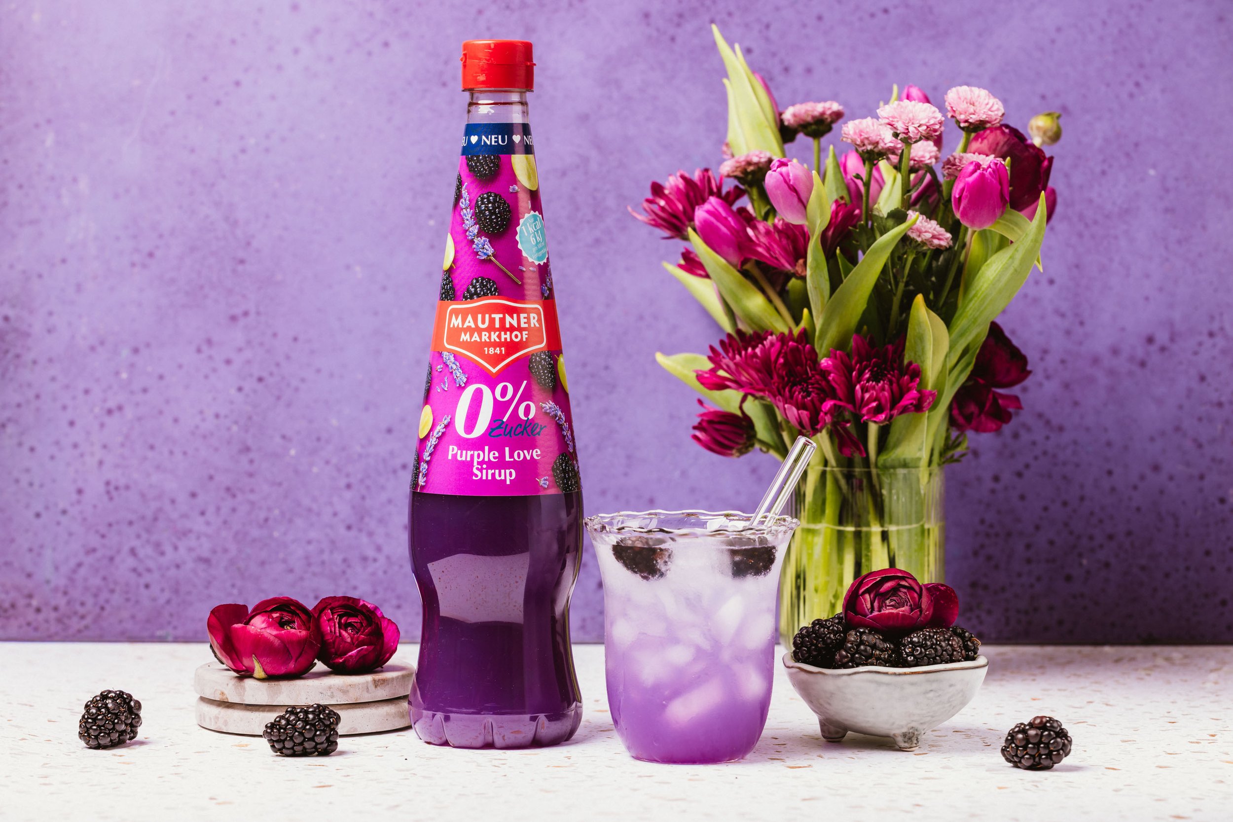 Ein Glas mit Blaubeeren, ein Flasche mit lavendelfarbigem Sirup, eine Blumenvasen mit pinken und lila Blumen, eine kleine Schüssel mit schwarzen Beeren, einige weiße Untersetzer, pinke Blüten und schwarze Beeren auf einem weißen Tisch vor einem viole