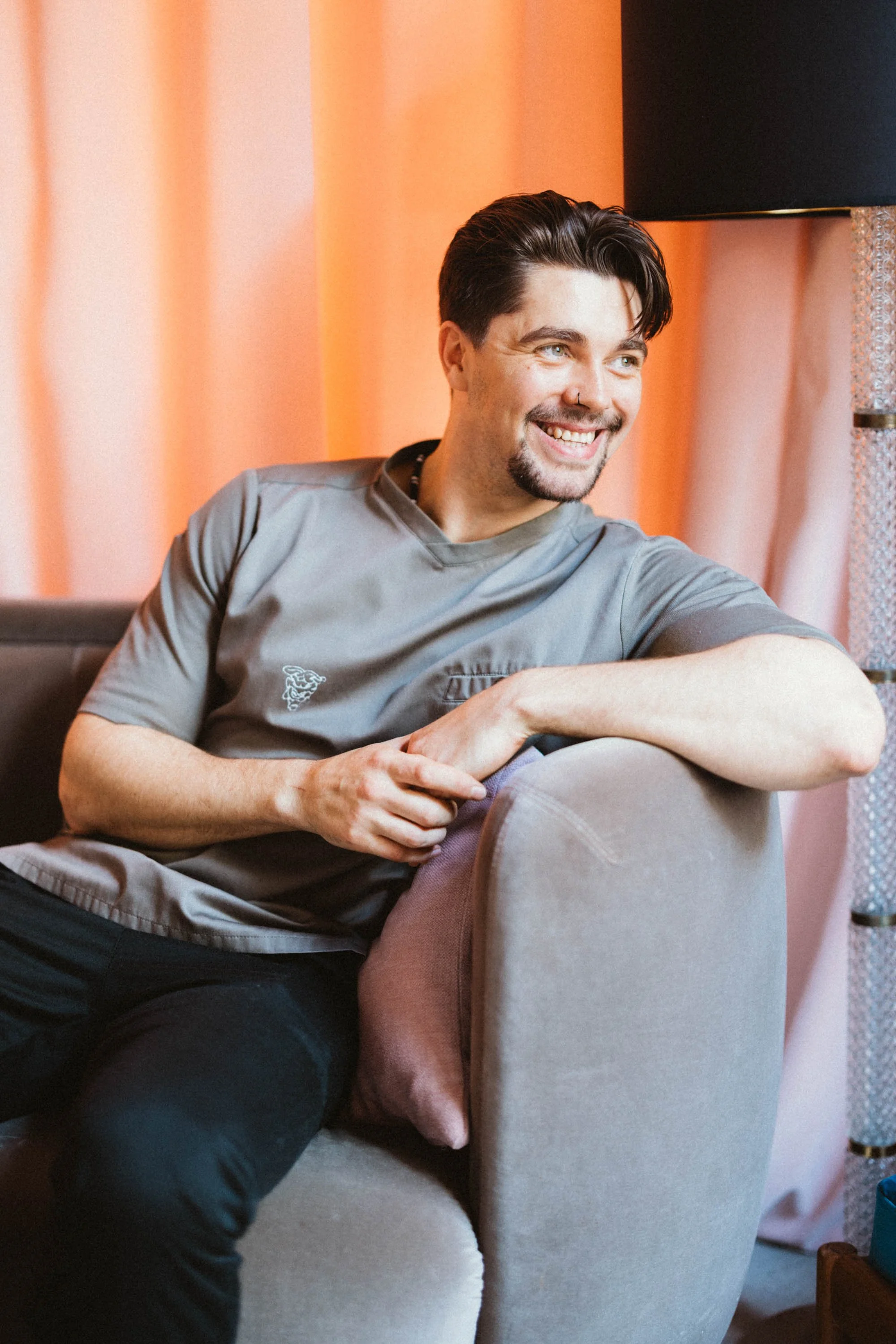 Ein lachender junger Mann mit dunklen Haaren und Brille sitzt auf einem grauen Sofa vor orangen Vorhängen.