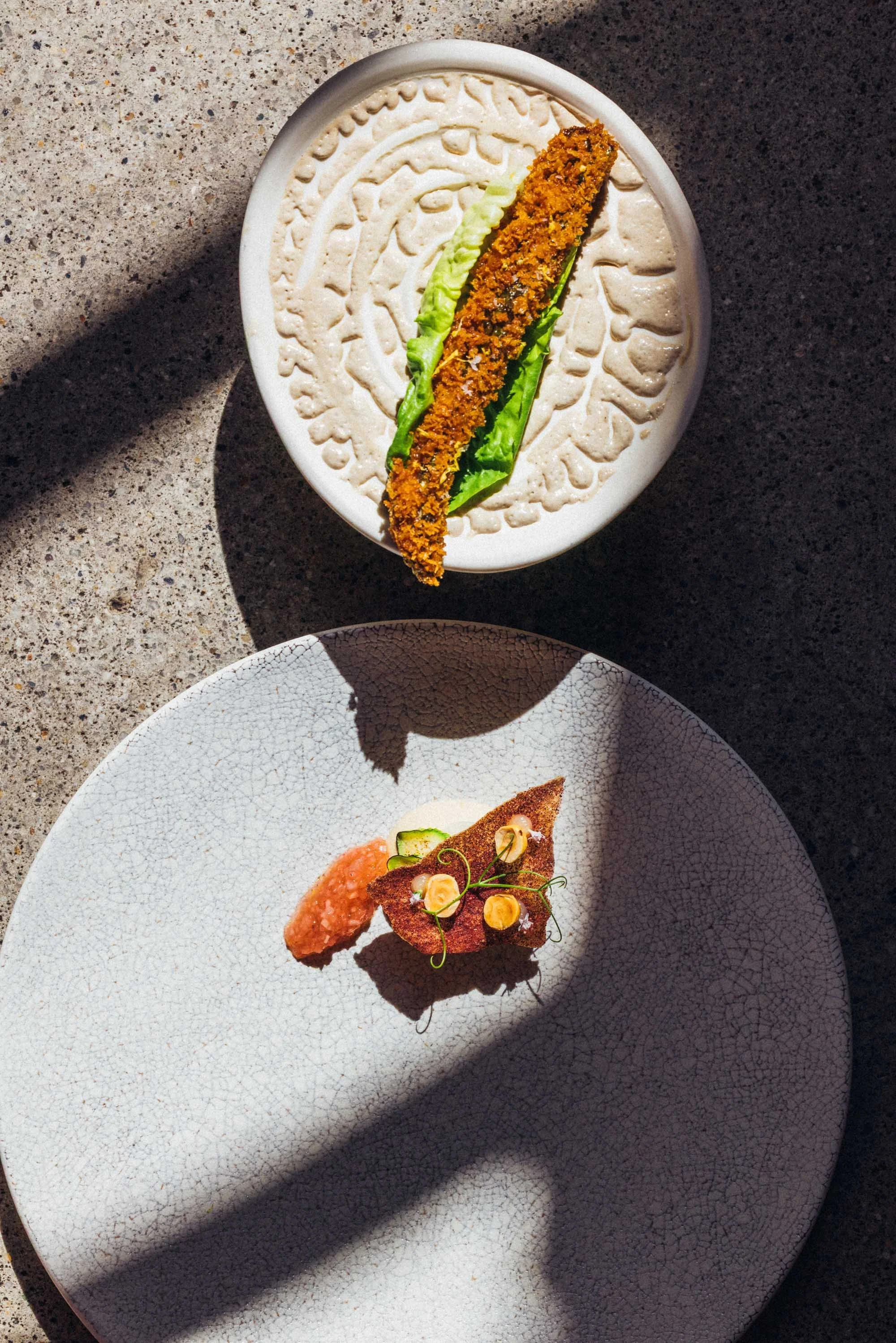 Zwei Teller mit kunstvoll angerichteten Speisen auf einer Terrasse. Der obere Teller hat ein Donut-artiges Gebäck mit grünen Salatblättern. Der untere Teller enthält ein kleines, dekoratives Gericht mit einer knusprigen braunen Basis, Gurkenscheiben 