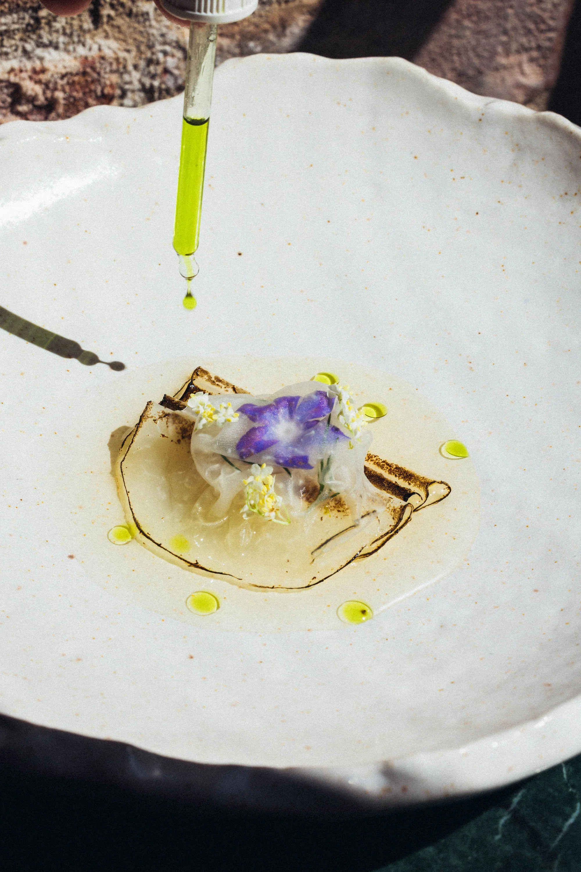 Feines Gericht auf weißem Teller mit blauer Blume, kleiner gelber Dekoration und dünner dunkler Linie, bei dem Öl mit Pipette darüber gereicht wird.