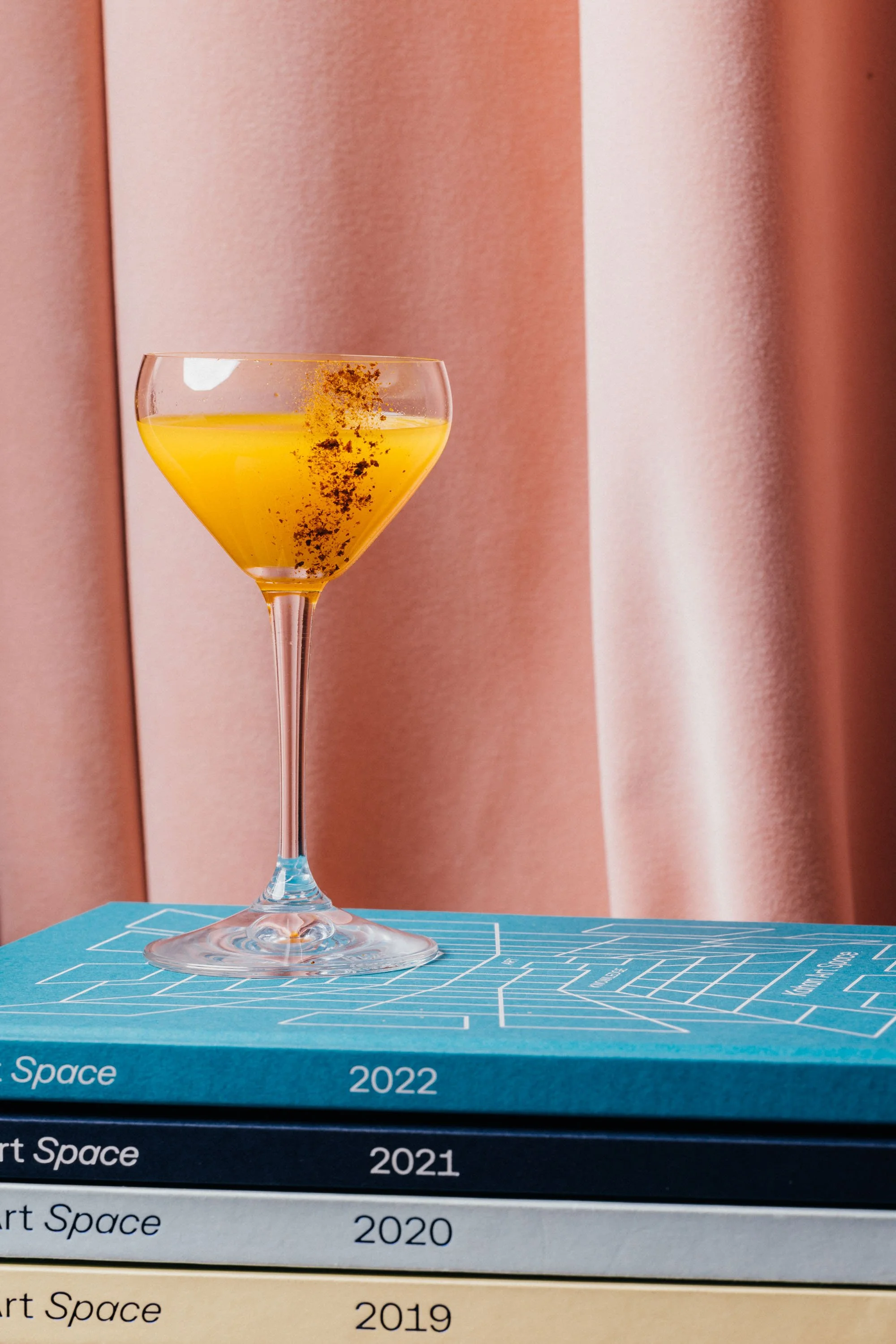 Ein Cocktails aus einem Martini-Glas mit gelber Flüssigkeit, dekoriert mit schwarzen Brocken, sitzt auf einer Blu-ray-Boxenregal. Im Hintergrund eine rosa-rosafarbene Stoffvorhang.