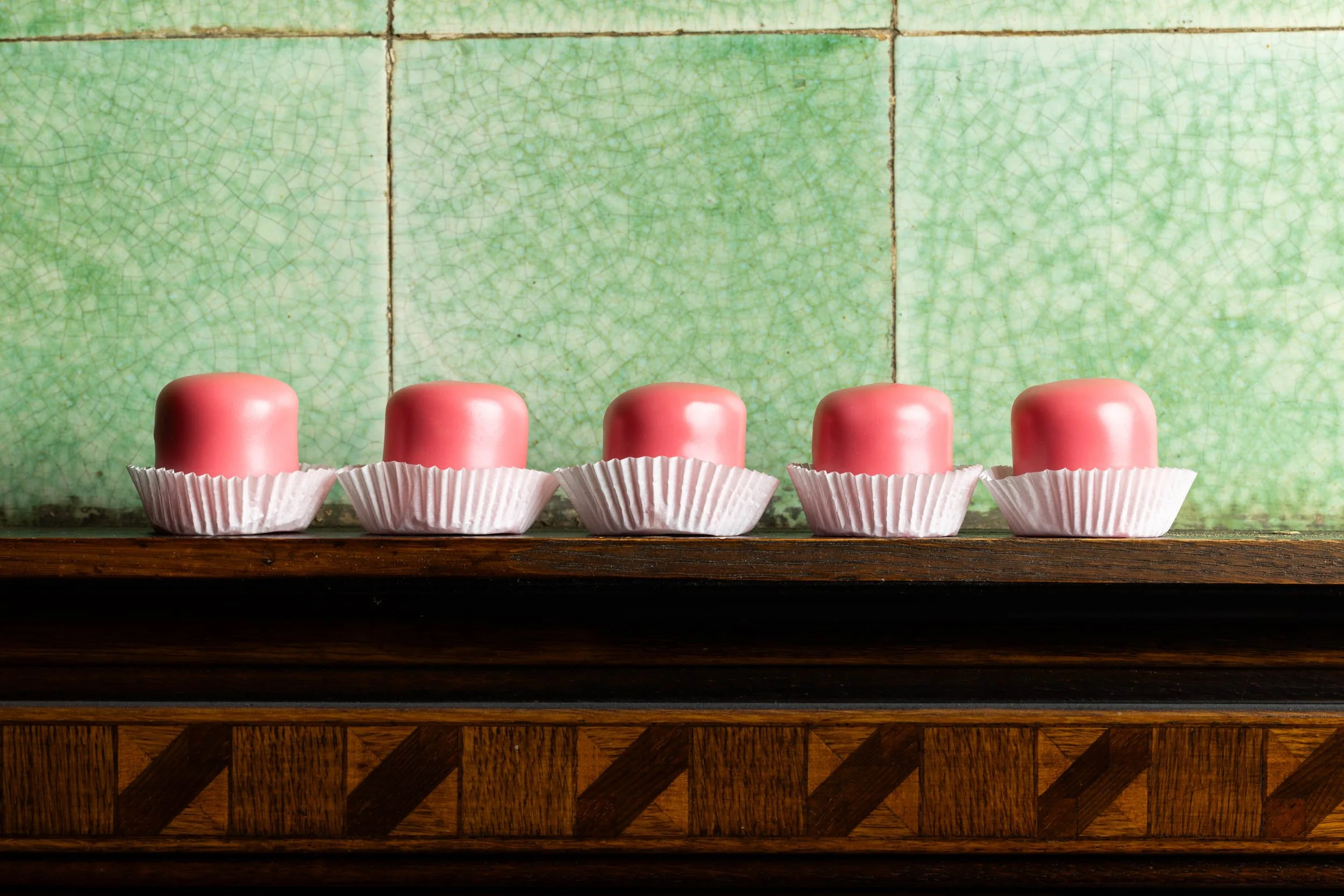 Fünf pinke Pralinen auf einer Holzplatte, jede in einem weißen Papierförmchen, vor einer grünen, gekachelten Wand.