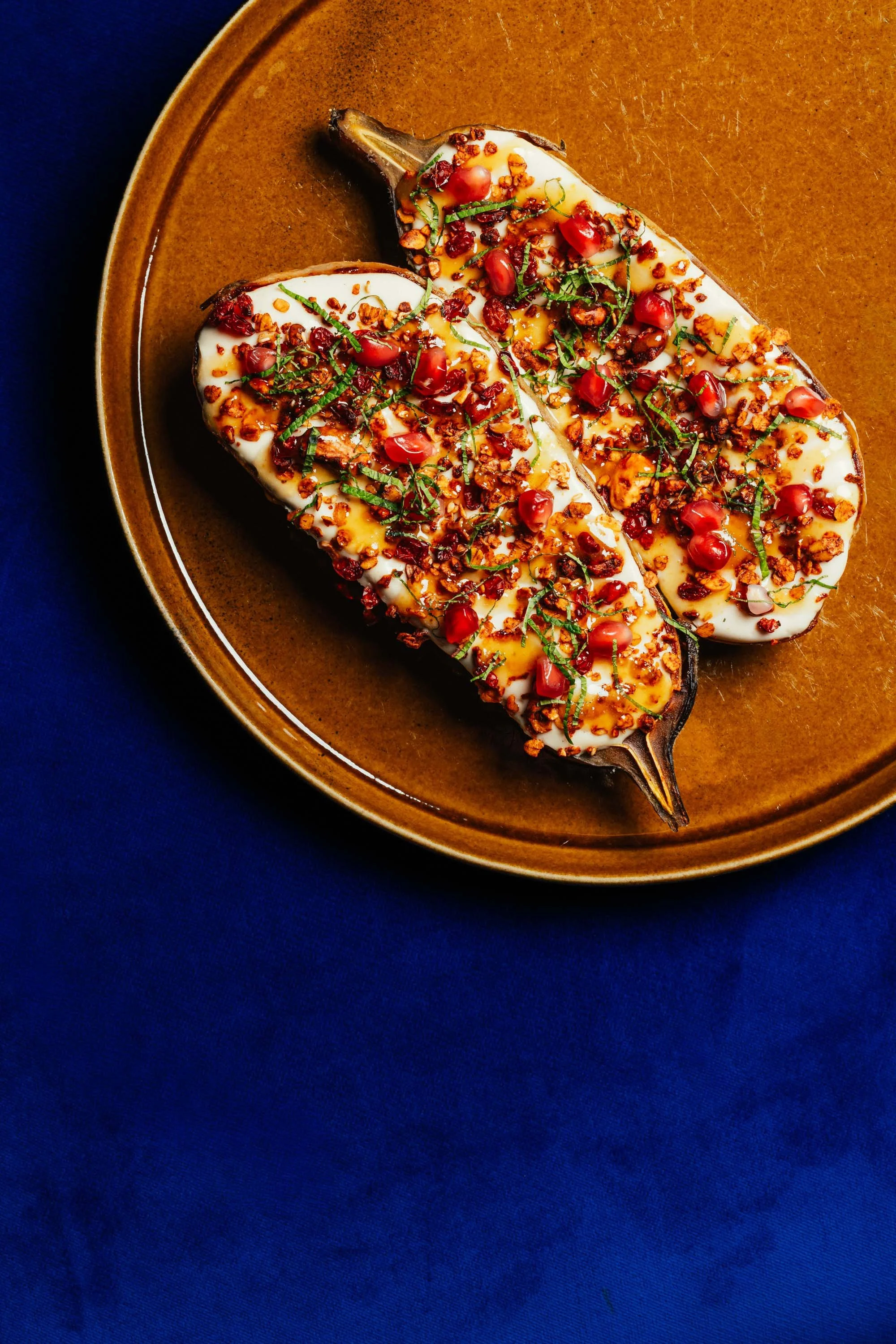 A grilled eggplant halved and topped with creamy sauce, pomegranate seeds, chopped herbs, and crispy crunchy topping on a brown plate.