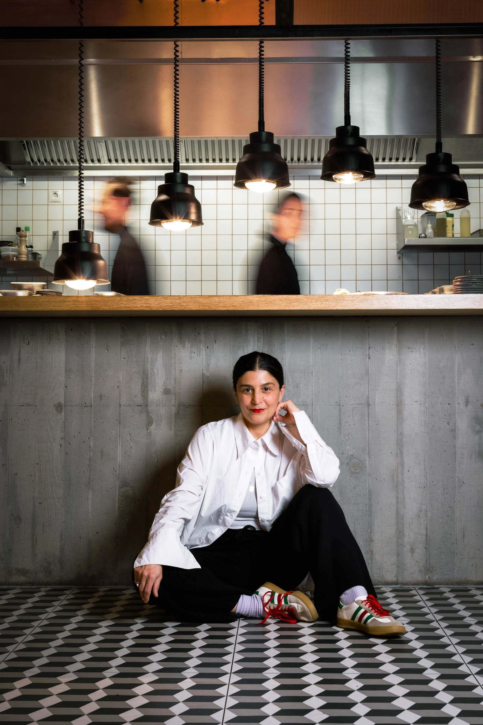 Eine Frau sitzt auf dem Boden vor einer grauen Wand, trägt weiße Hemden, schwarze Hosen und Sneakers, während im Hintergrund die Küchenmitarbeiter beim Kochen sichtbar sind.