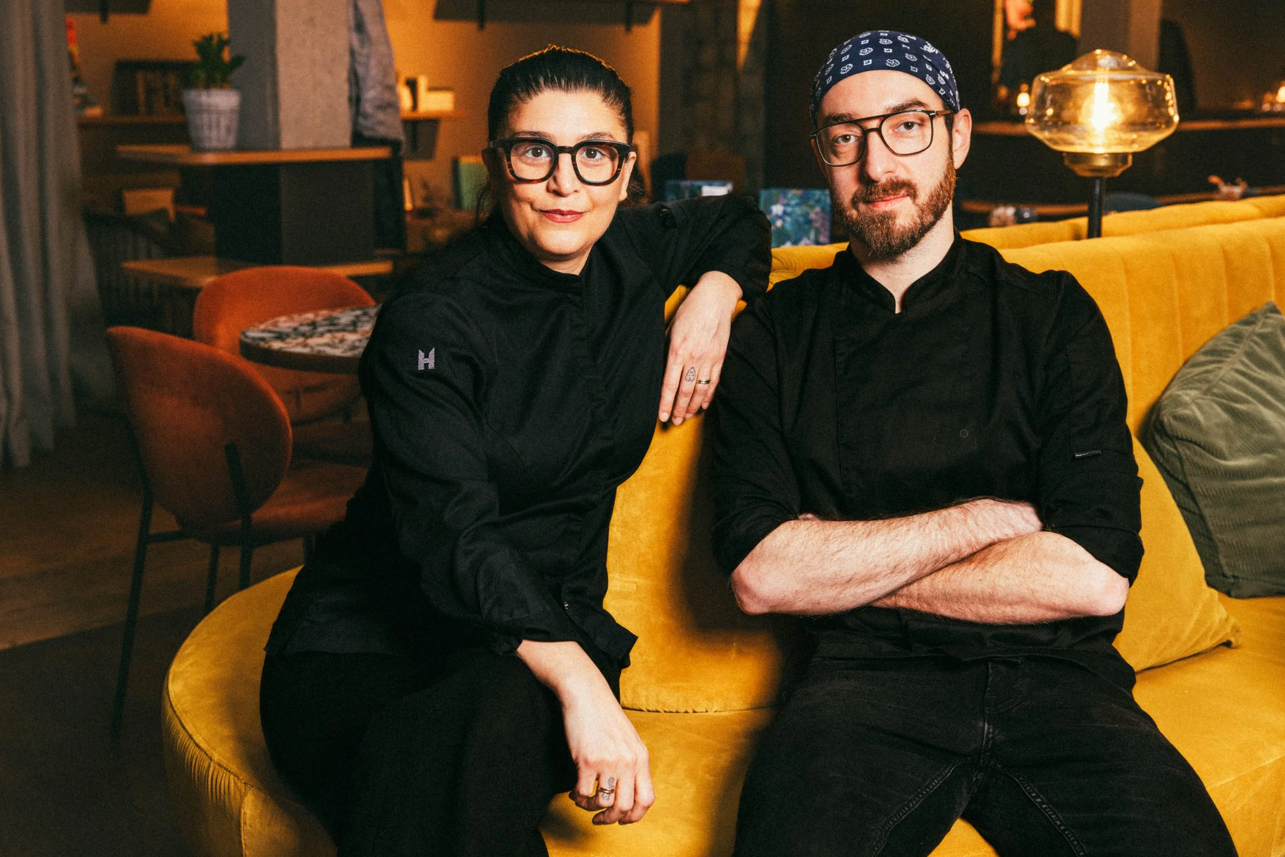 Zwei Köche sitzen auf einem gelben Sofa in einem eleganten Restaurant, einer mit hochgestecktem Haar und Brille, der andere mit Bart, Brille und Bandana.