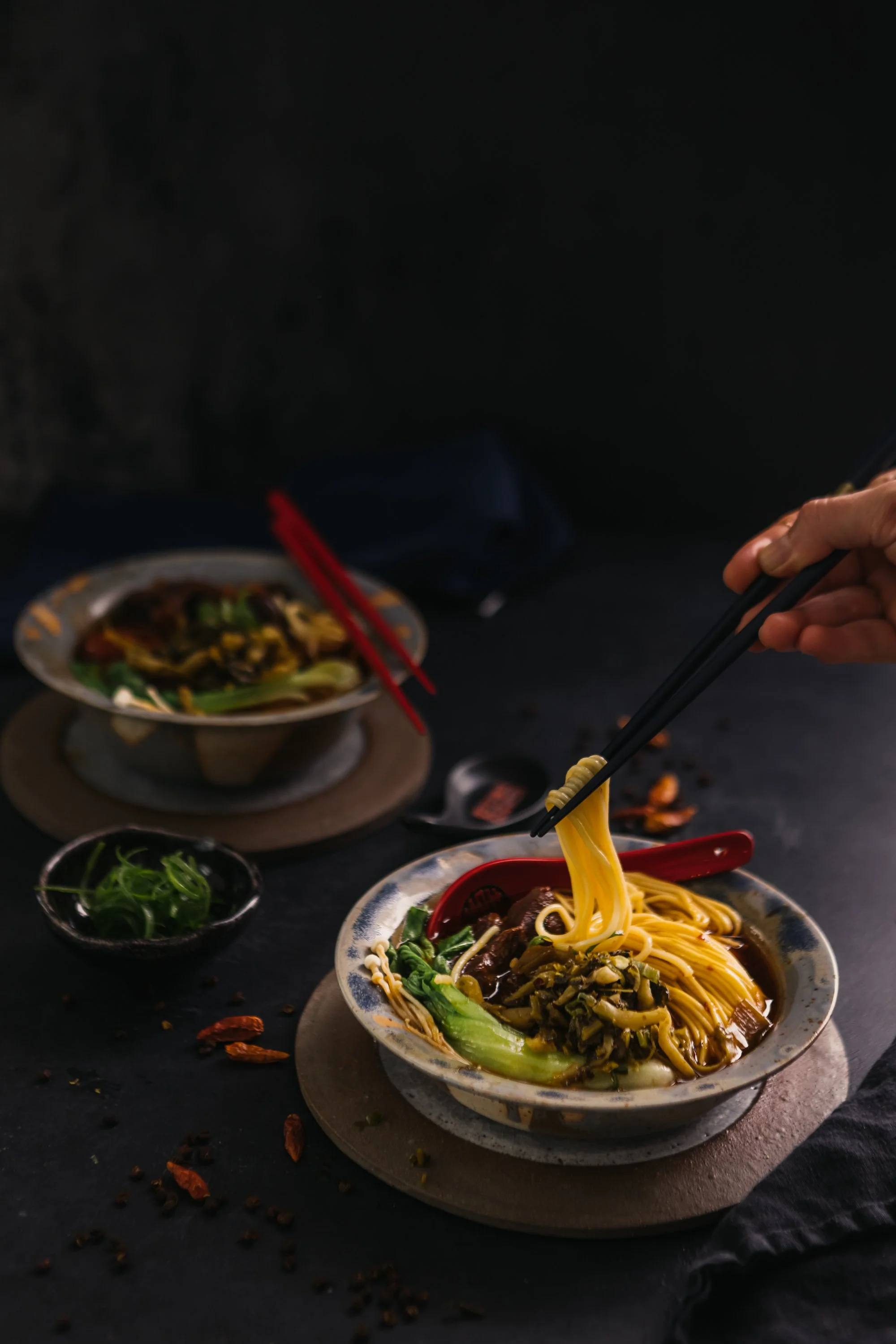 Nahaufnahme von zwei Schüsseln mit asiatischem Menü, eine mit Nudeln und Gemüse, eine andere im Hintergrund, auf einem schwarzen Tisch mit roten Essstäbchen und kleiner Schüssel mit grünen Kräutern, dunkler Hintergrund.