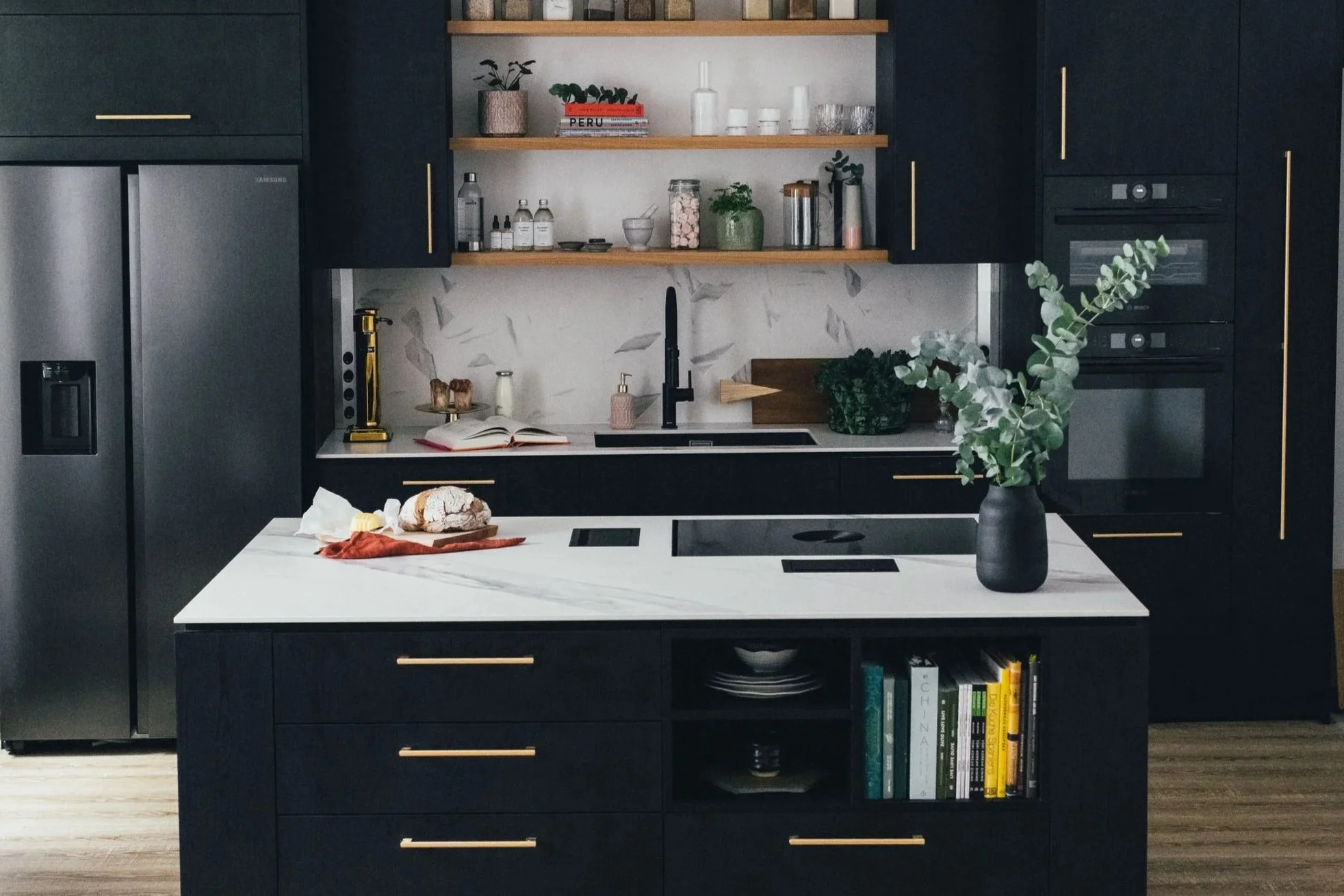 Moderne schwarze Küche mit weißer Marmor-Arbeitsfläche, offenen Holzregalen, verschiedener Küchenutensilien, schwarzen Geräten und einem großen Kücheninsel, die mit einem schwarzen Vase mit grünen Zweigen dekoriert ist.