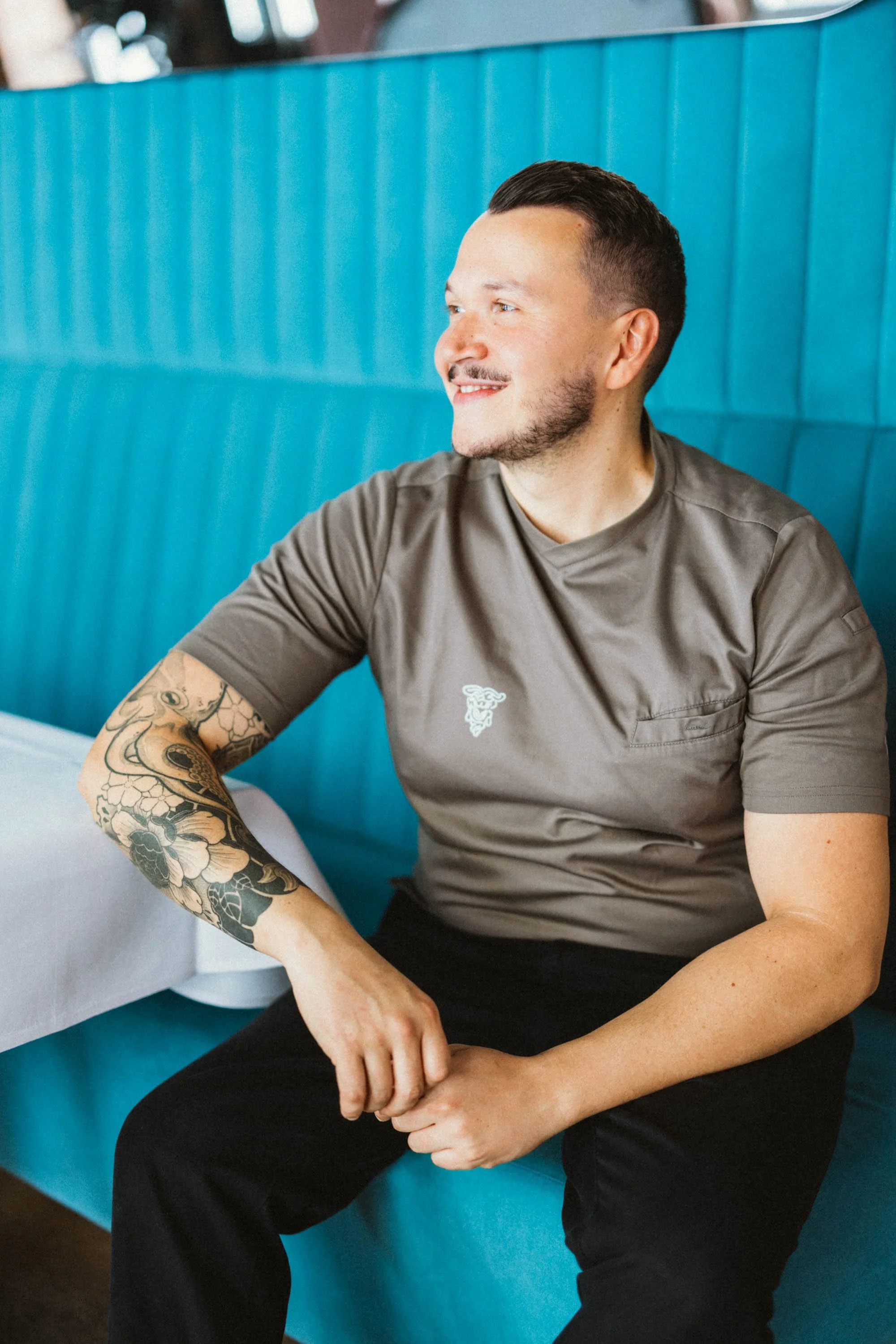 Ein lachender Mann mit Tattoos sitzt in einem Restaurant, trägt ein braunes T-Shirt und schwarze Hosen, vor einem blauen mit Polstern versehenen Wand.
