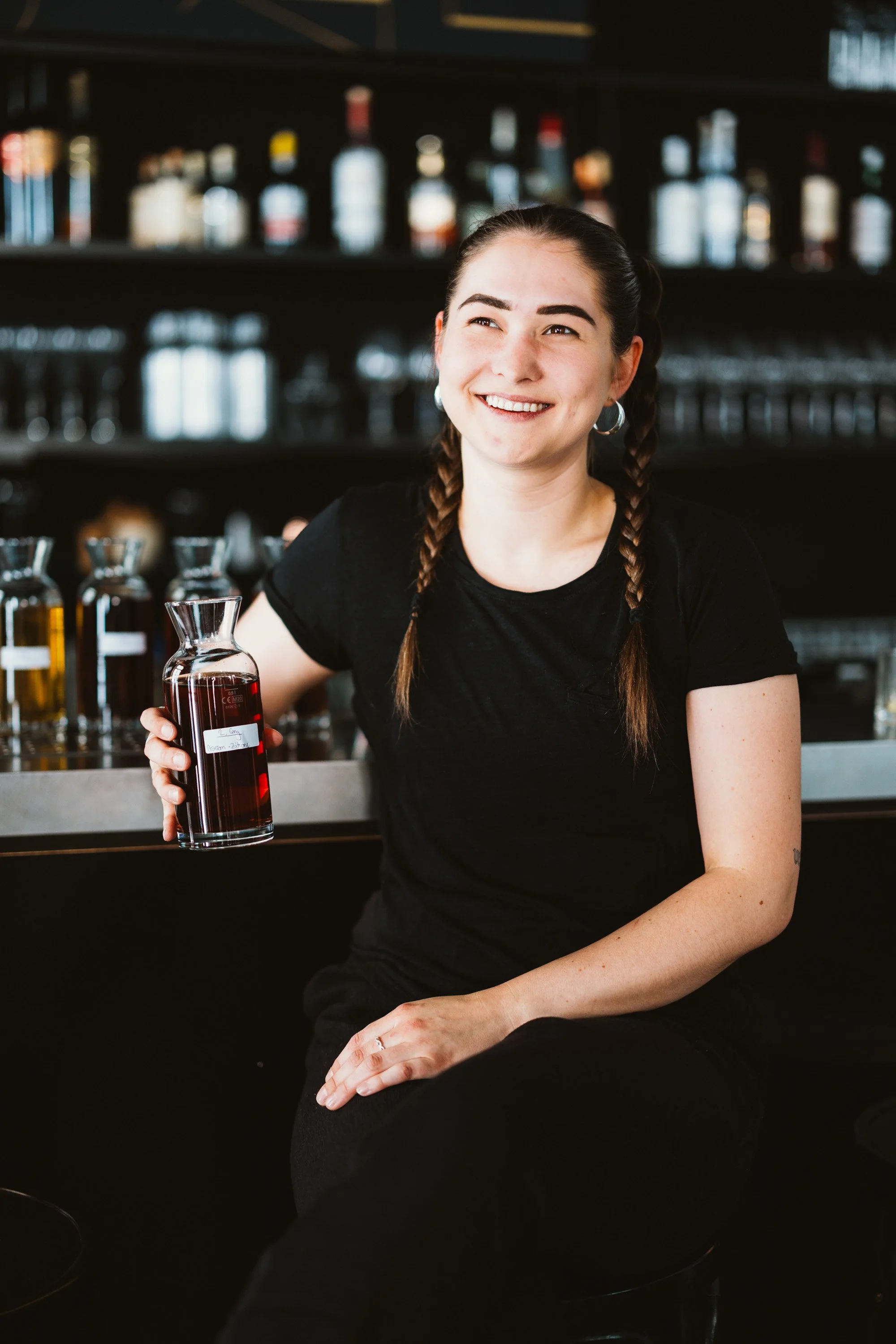 Junge Frau mit Zöpfen und schwarzen Kleidung sitzt an einer Bar und hält eine Flasche mit rotem Getränk.