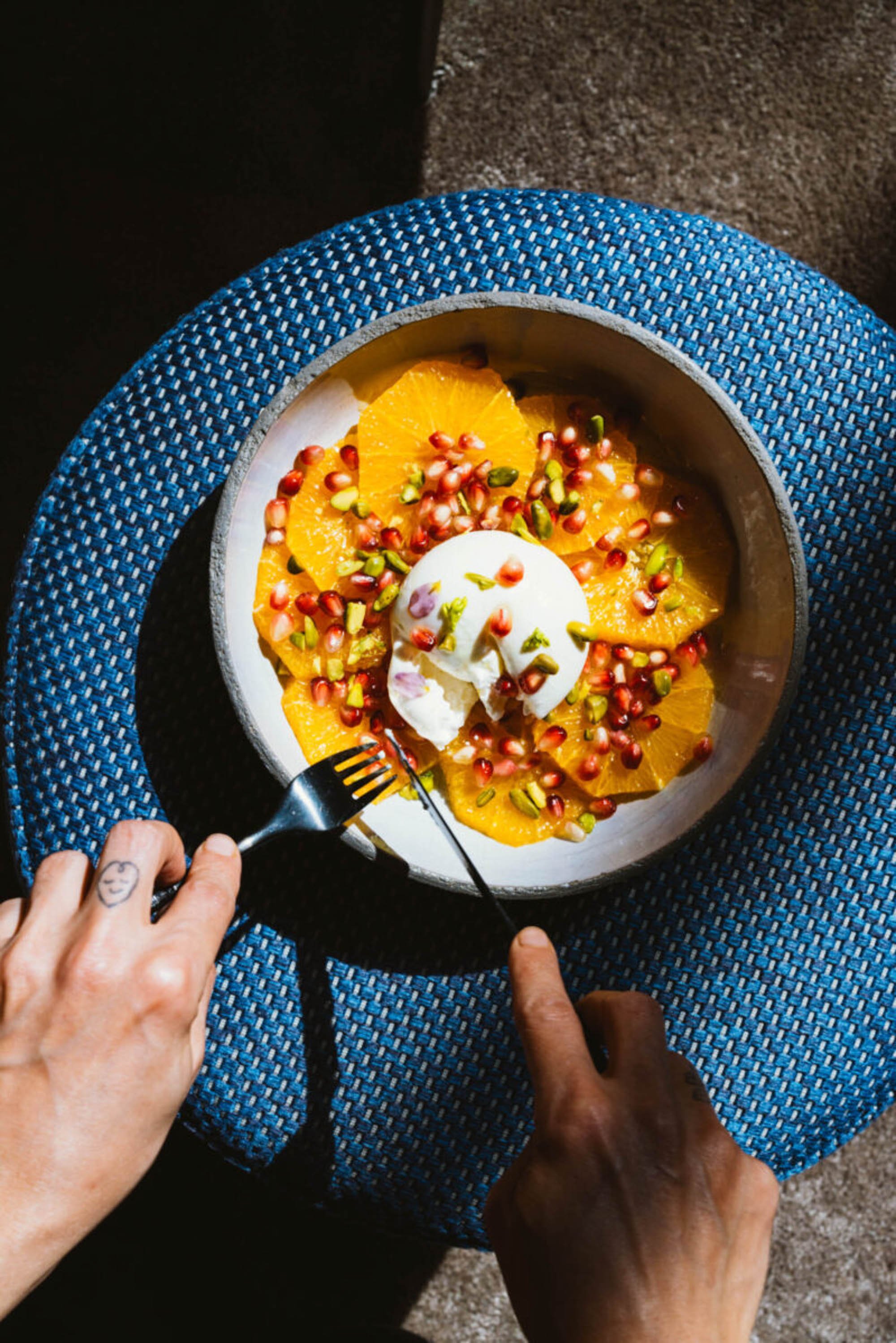 Schüssel mit Orangenscheiben, Joghurt, Granatapfelkerne und Pistazien auf einem blauen Tisch. 