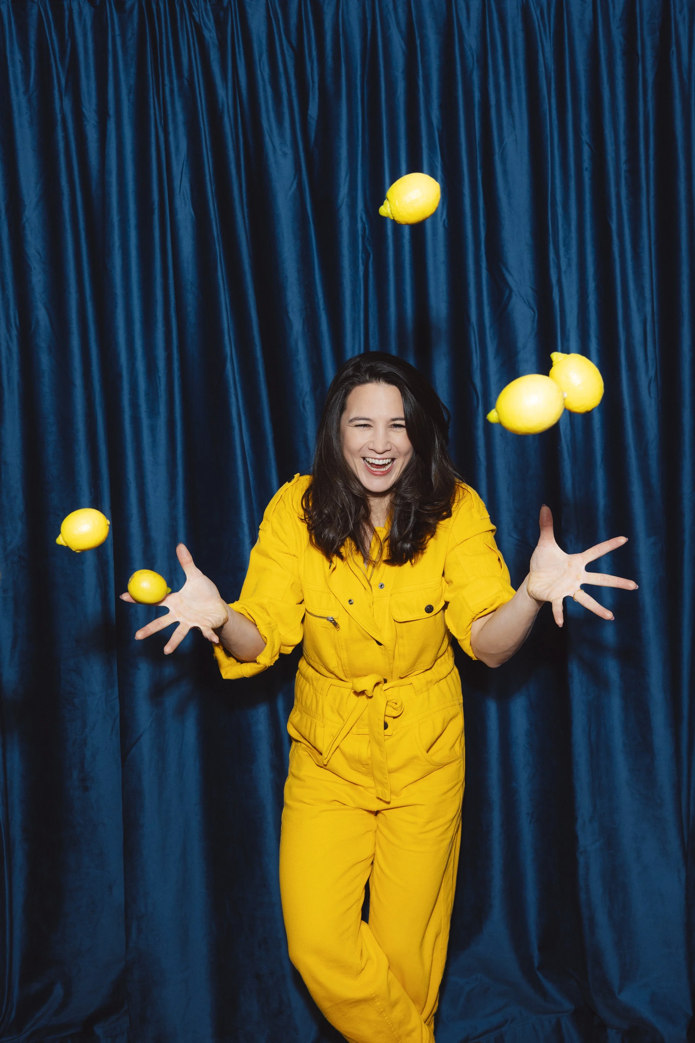 Lächelnde Frau in gelbem Anzug jongliert mit gelben Zitronen vor dunklem blauen Vorhang.