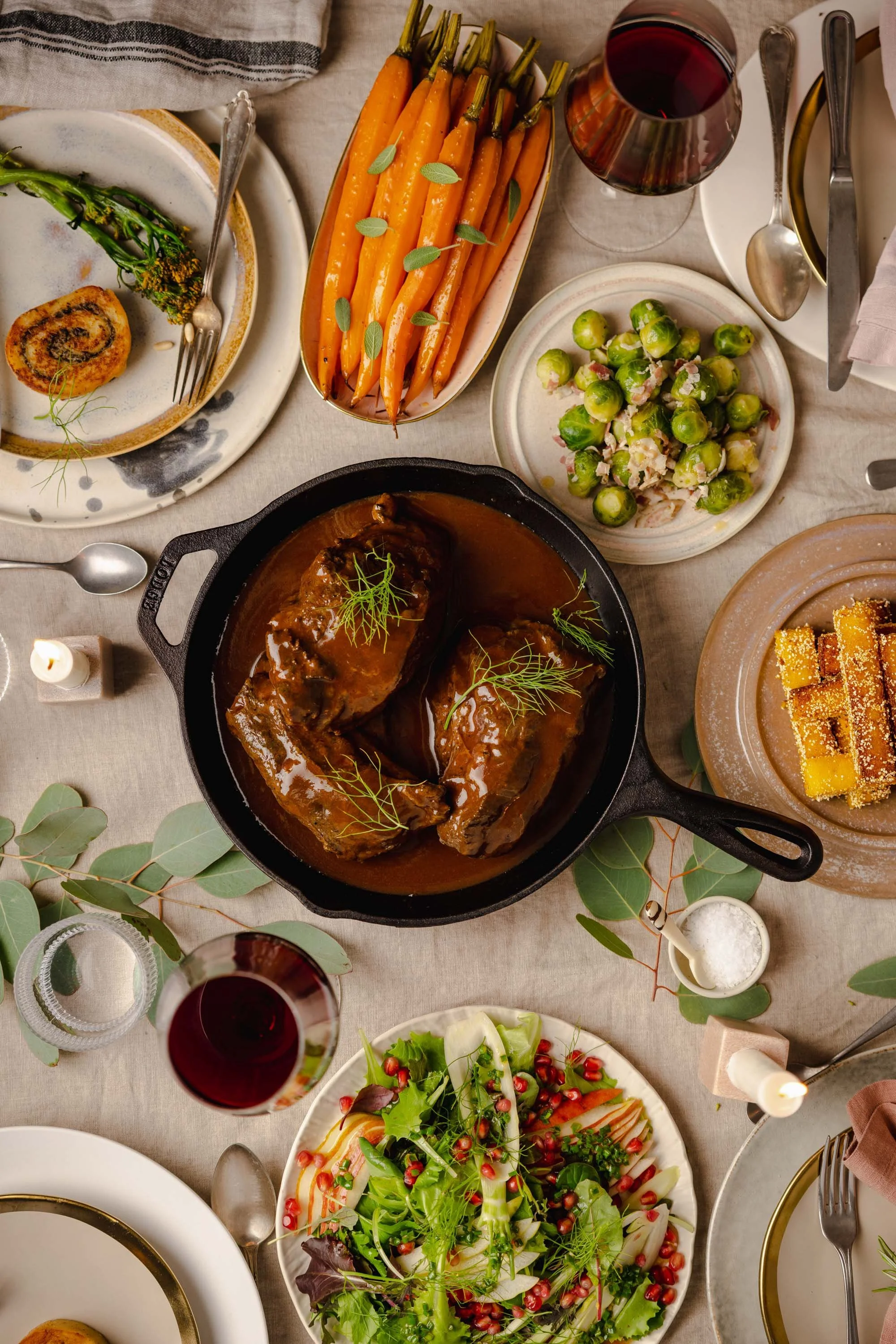 Ein gedeckter Tisch mit verschiedenen Gerichten, darunter eine Fleischportion in Sauce, ein Salat mit Granatapfelkernen, geröstete Karotten, eine gebackene Rolle, eine Gemüsesalle, ein Glas Rotwein und ein Dessert mit gelben Stückchen.