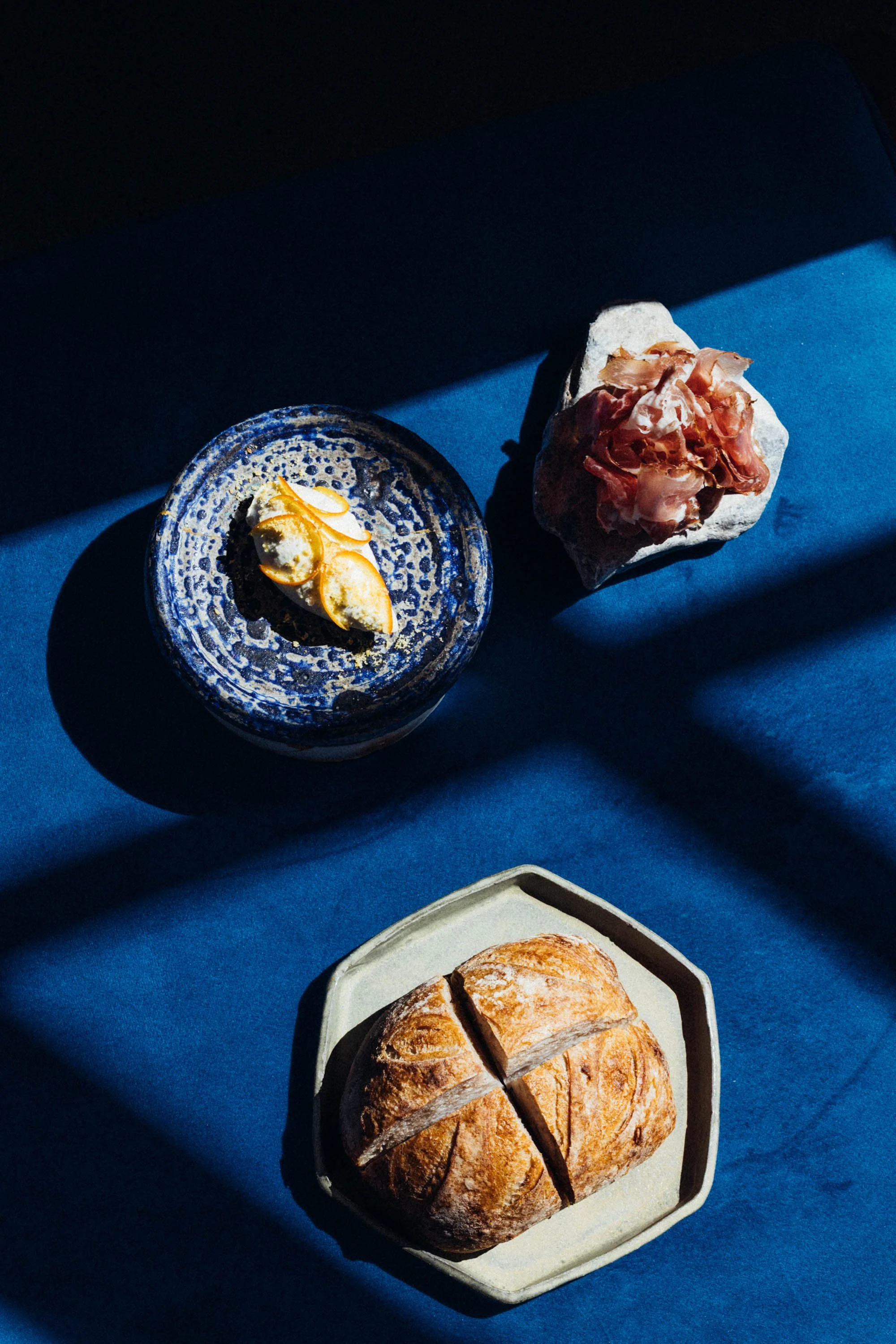 Brot, Schinken, Käse und eine kleine Schale auf einem blauen Tuch im Sonnenlicht.