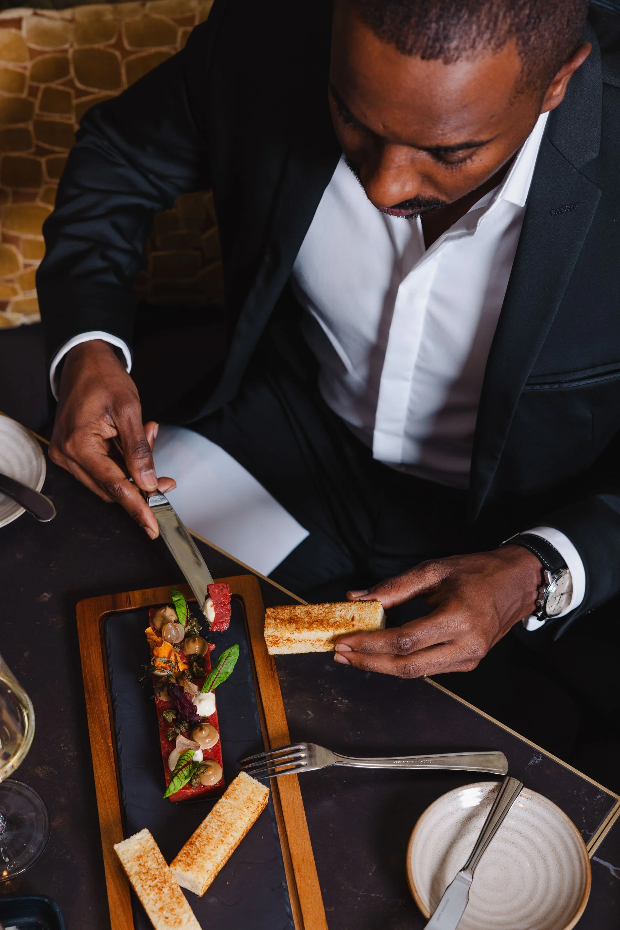 Ein Mann in einem schwarzen Anzug schneidet ein Gericht mit einer Gabel und einem Messer, das auf einem schwarzen Teller serviert wird, der auf einem Holzbrett liegt, während er an einem gedeckten Esstisch sitzt.