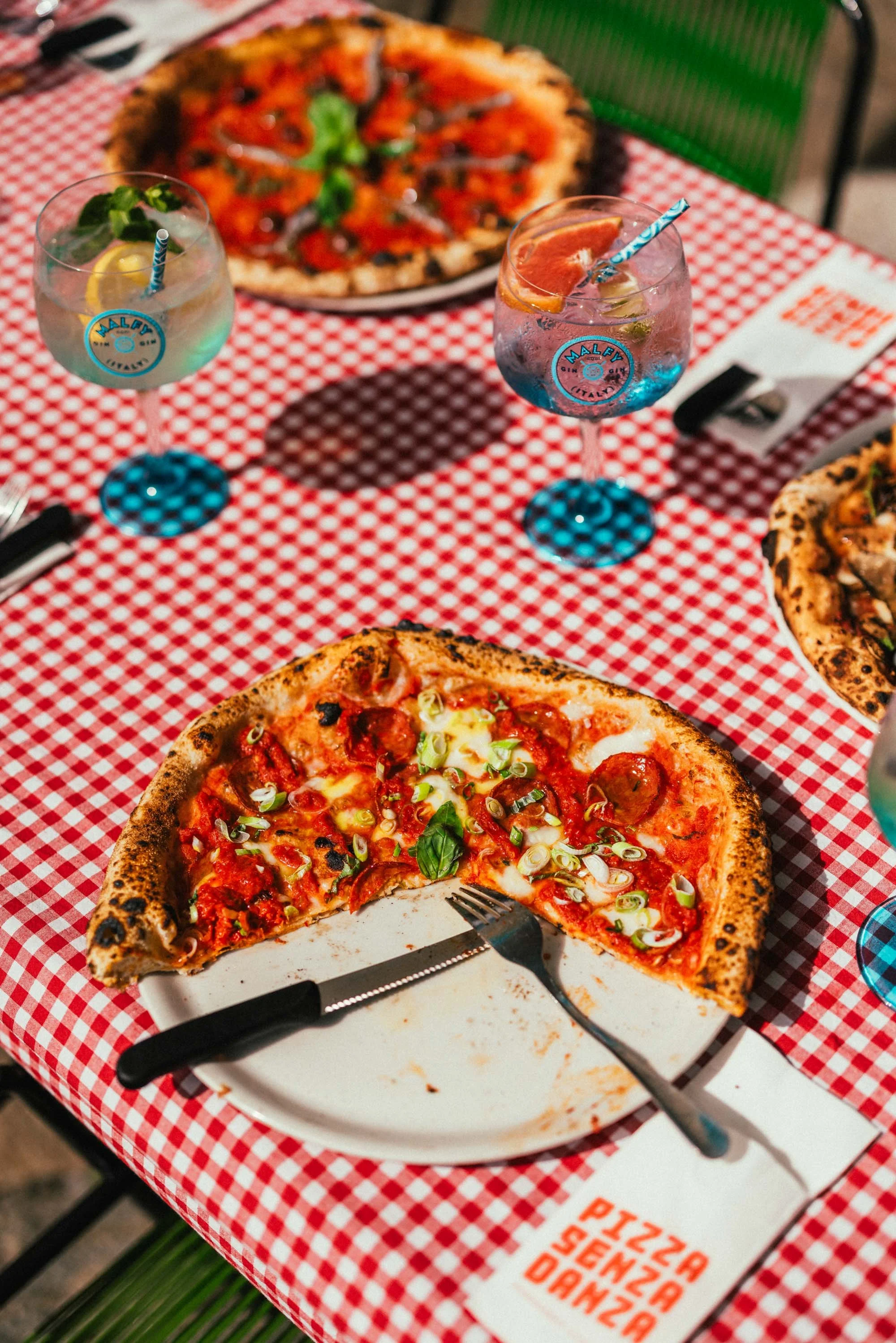 Eine halbe Pizza auf einem weißen Teller, auf einem rot-weiß karierter Tischtuch, mit einem Messer und einer Gabel. Im Hintergrund sind zwei bunte Getränke in Gläsern, jeweils mit einer Zitronen- und Limettenscheibe sowie einem Strohhalm. Der Tisch h