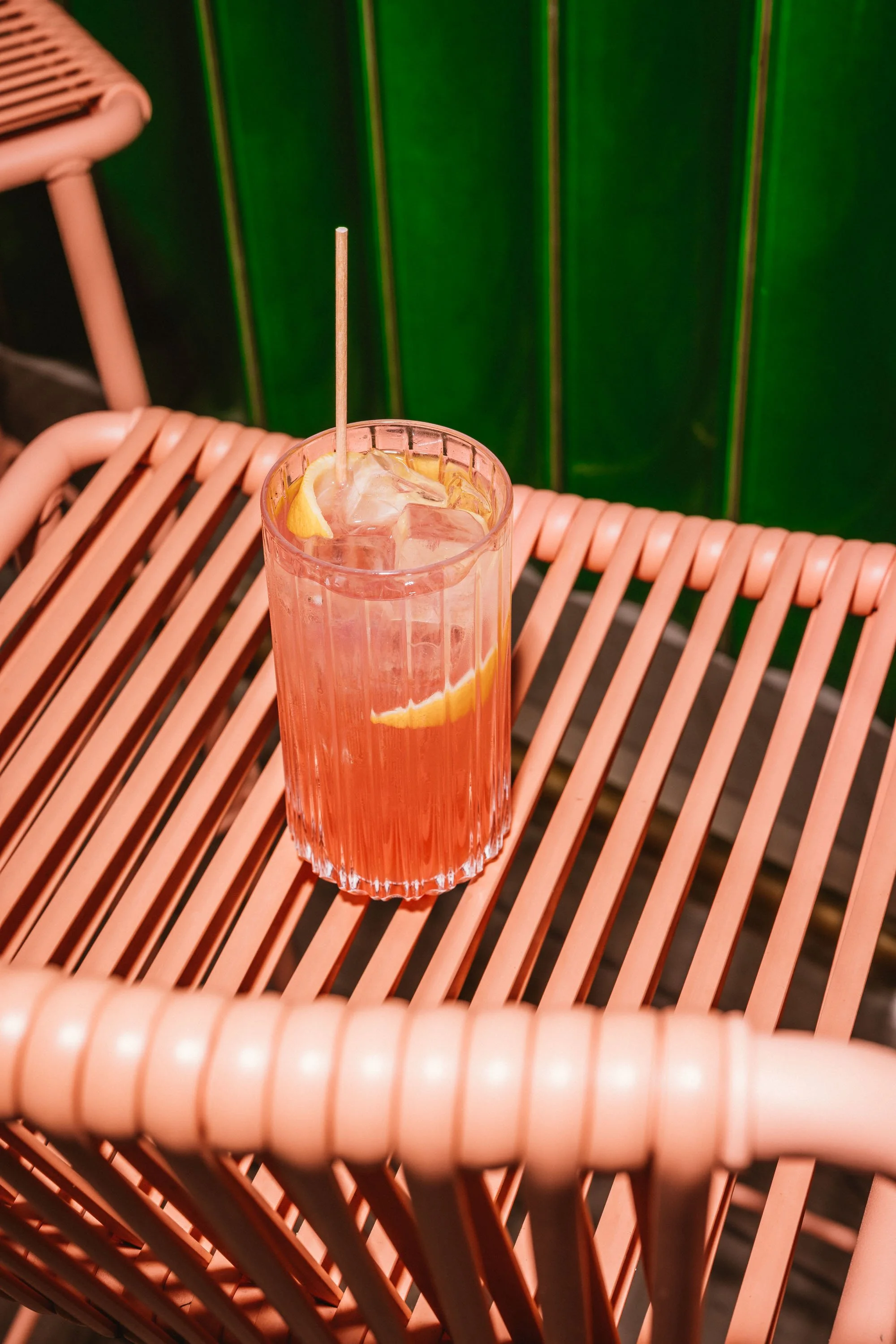 Ein Glas mit rosa Getränk, Zitronenscheiben und Eis auf einem pinken Rattantisch