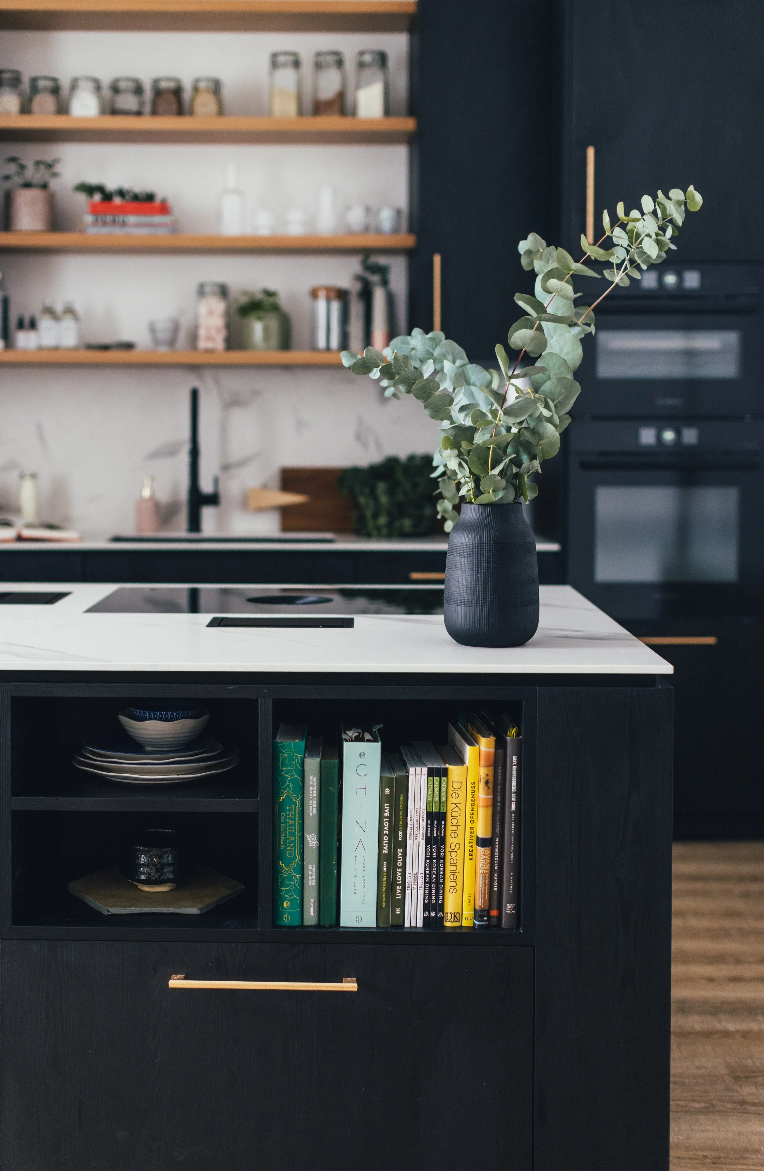 Schwarzes Küchenregal mit Bücher, darunter eine Vase mit Eukalyptus auf einer weißen Kücheninsel, im Hintergrund offene Holzregale mit Küchenutensilien und Dekorationen.