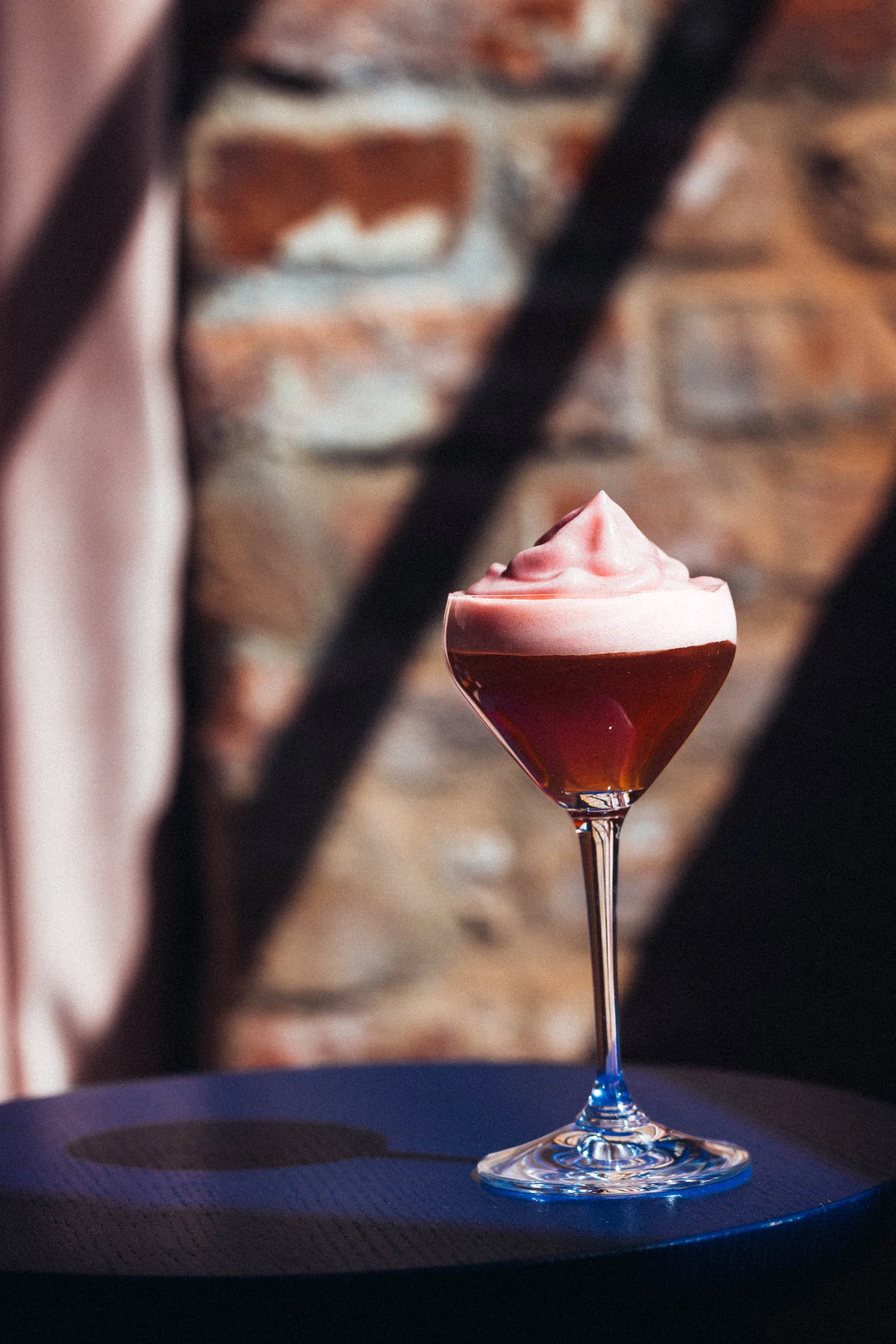 Ein Cocktail in einem Martini-Glas mit einem rosa Schaum oben, auf einem dunklen Tisch vor einer Ziegelsteinwand.