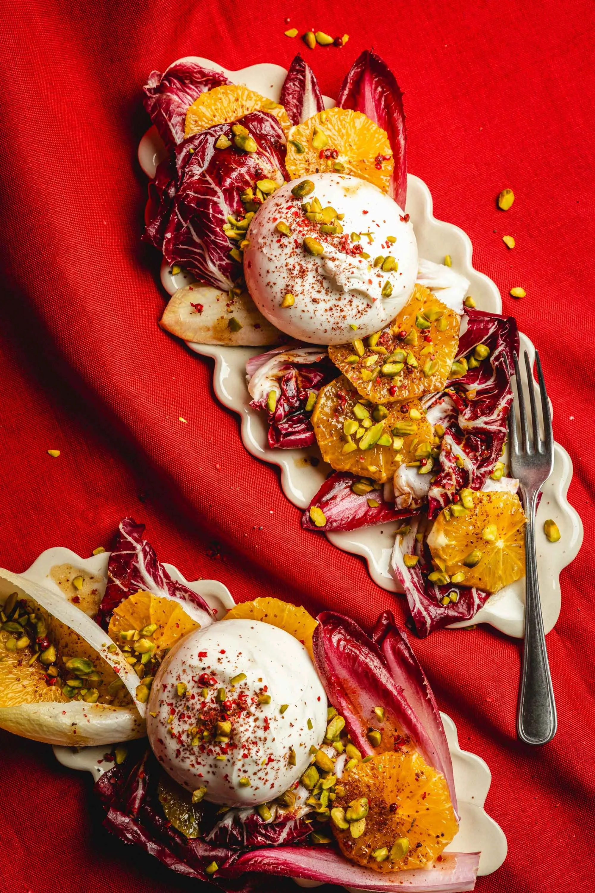 Zwei Teller mit Radicchio, orangen, Burrata-Käse, Pistazien und rotem Pfeffer auf rotem Tischtuch.