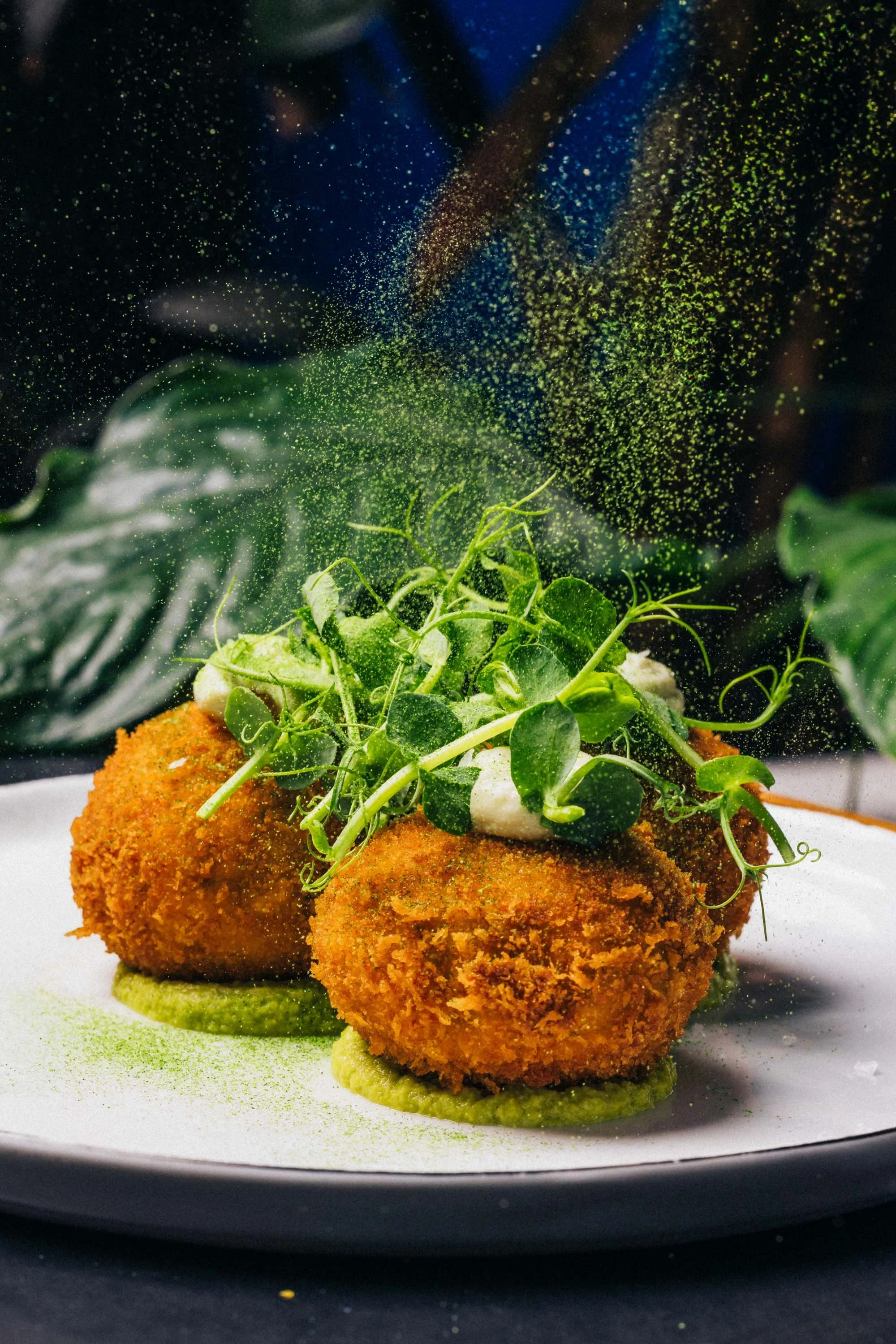 Frittierte Bällchen mit grüner Kräutern/Gemüse Garnitur und grüner Soße auf einem weißen Teller, mit Pulverstaub in der Luft.