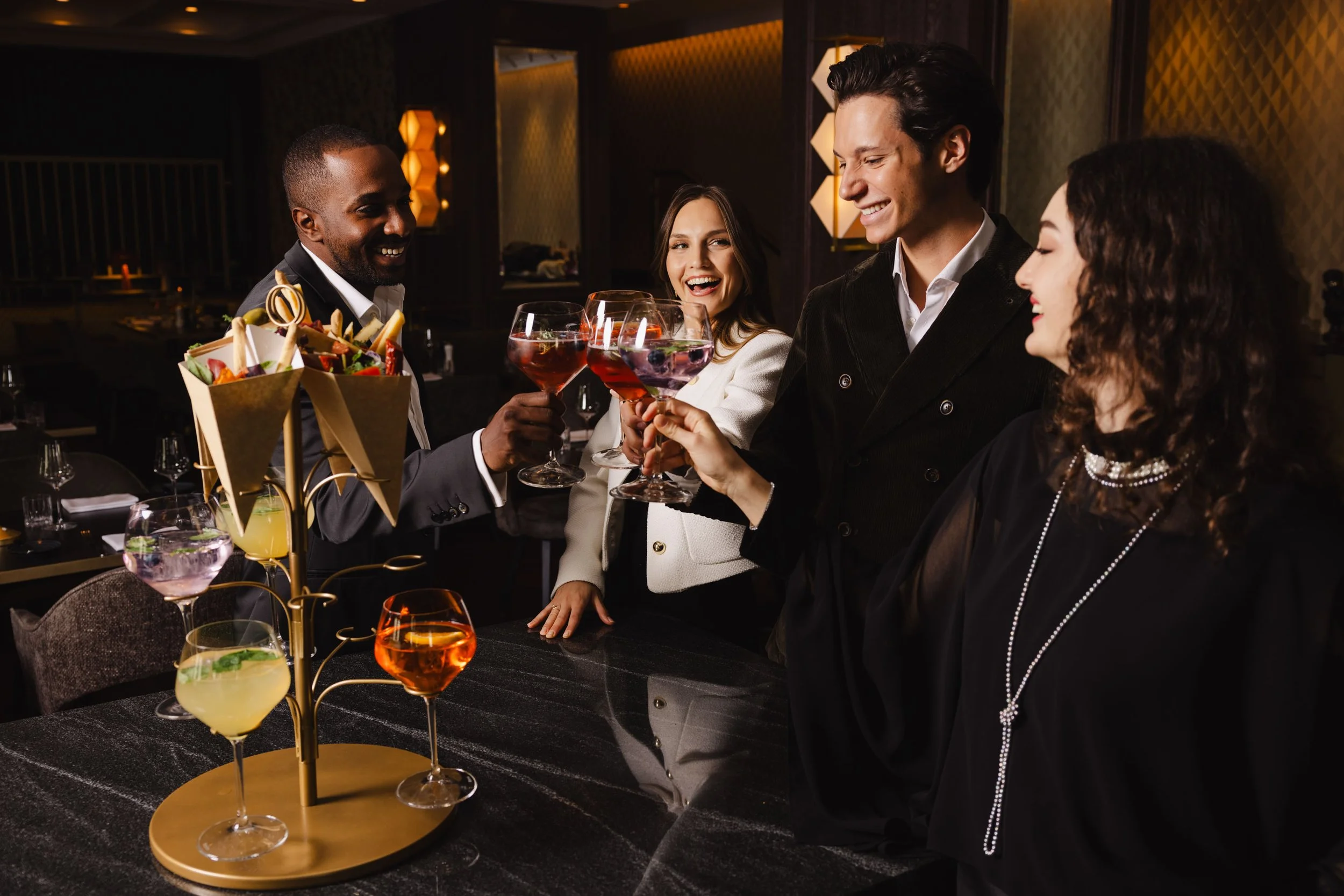 Menschen beim Anstoßen mit roten und gelben Getränken in einem eleganten Restaurant, mit einer dekorativen Obstplatte auf dem Tisch.