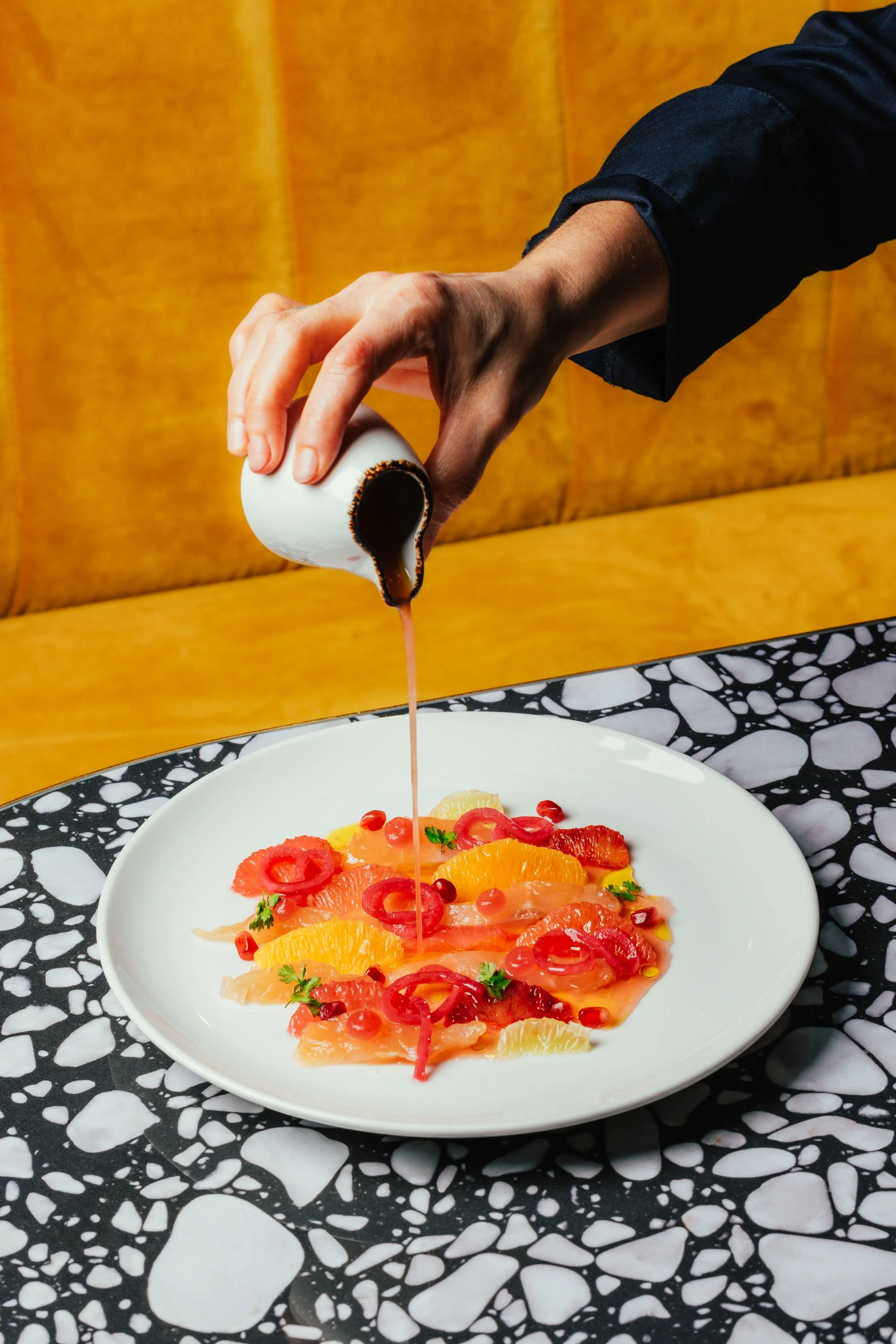Person gießt Sauce über einen Teller mit Früchten, darunter Orangen, Grapefruits und rote Beeren.