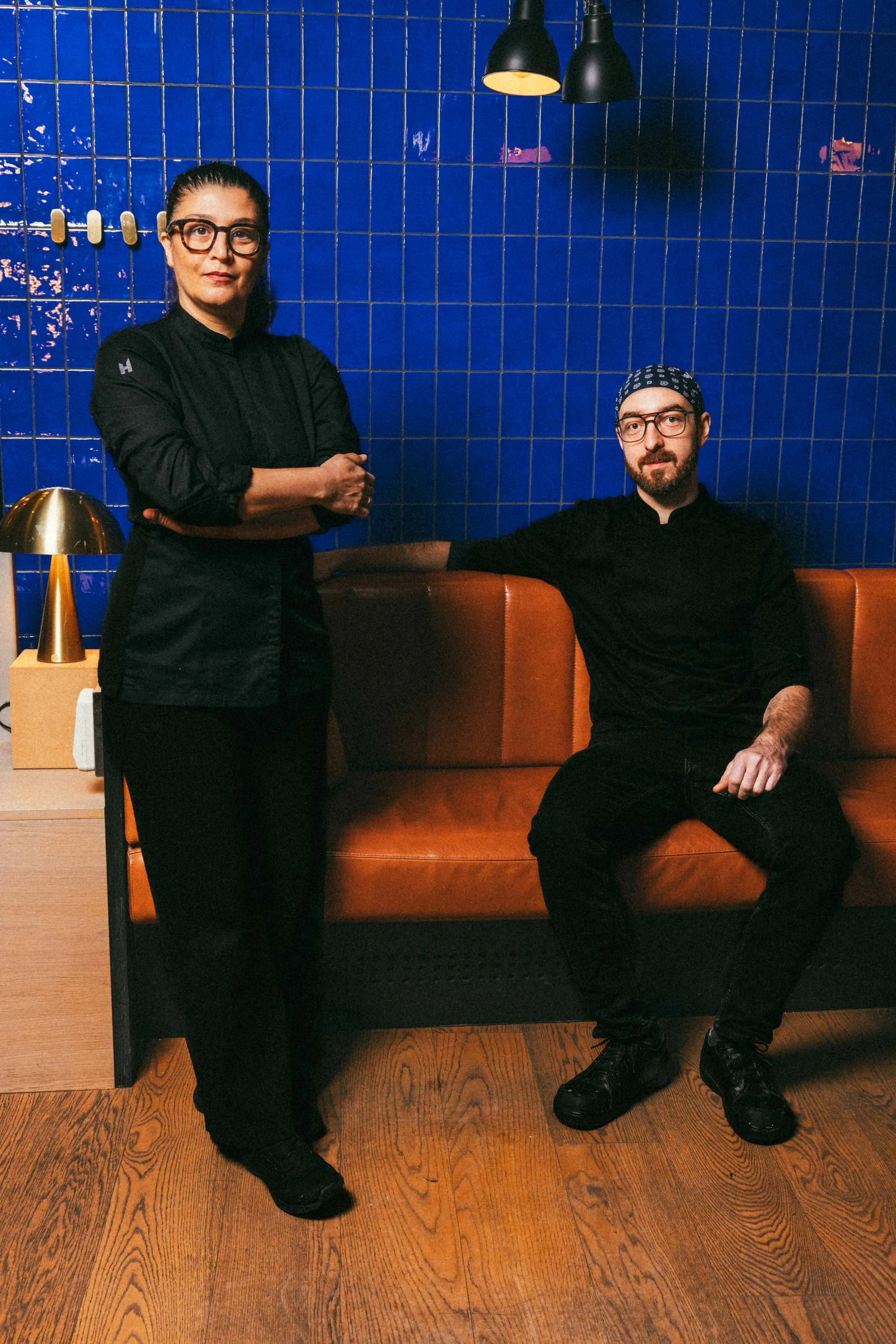 Zwei Köche in dunkler Kleidung, eine Frau stehend mit verschränkten Armen, ein Mann sitzend auf einer braunen Ledercouch vor einer blauen Fliesentapete, helles Licht von Deckenlampen, seitlich ein Nachttisch mit einem goldfarbenen Lampenschirm.