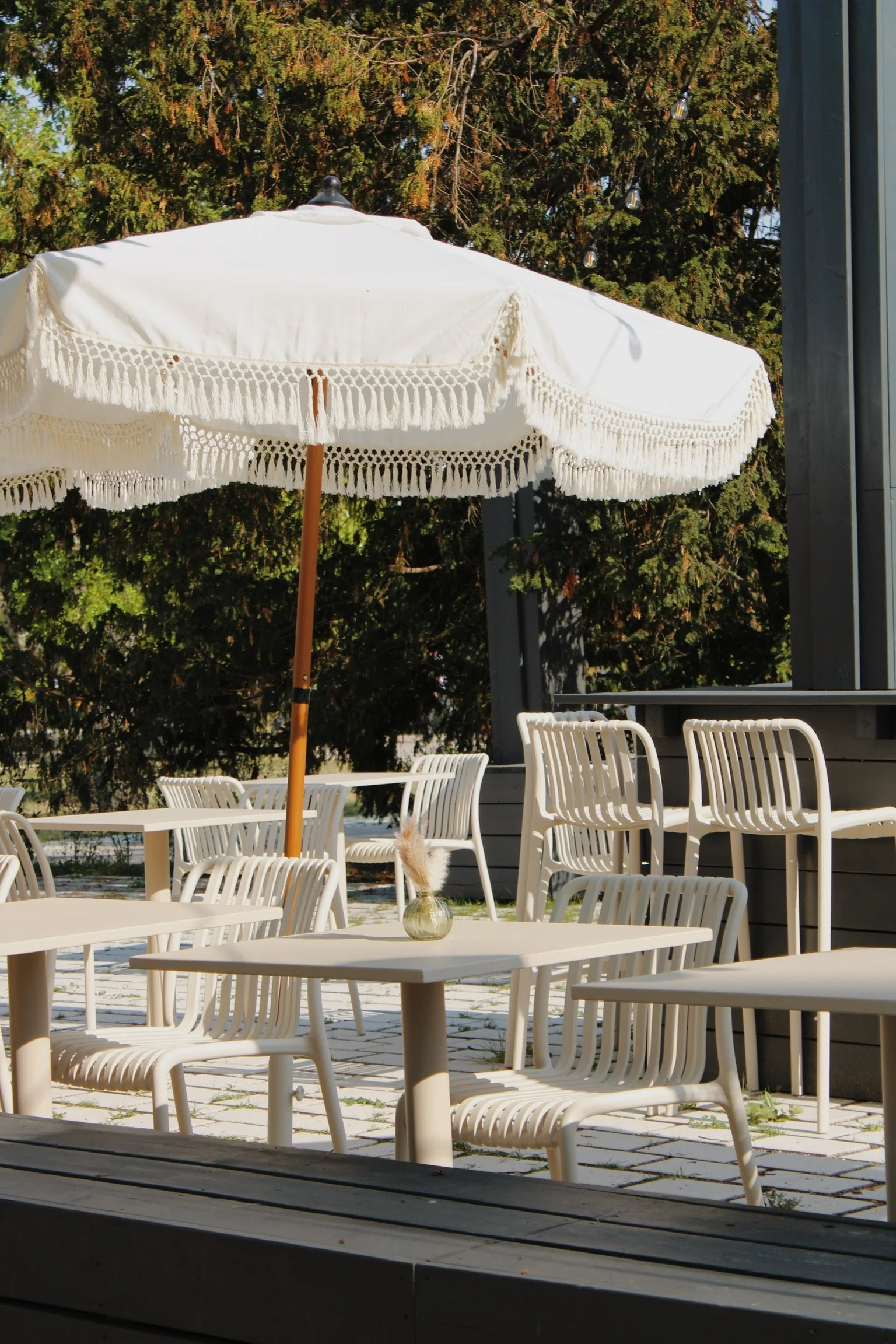 Terrasse extérieure avec parasol blanc en tissu ornée de franges, plusieurs chaises en plastique blanc autour de tables, un vase avec des fleurs séchées en premier plan, fond d'arbres verts, partie latérale en métal ou bois d'une structure moderne.