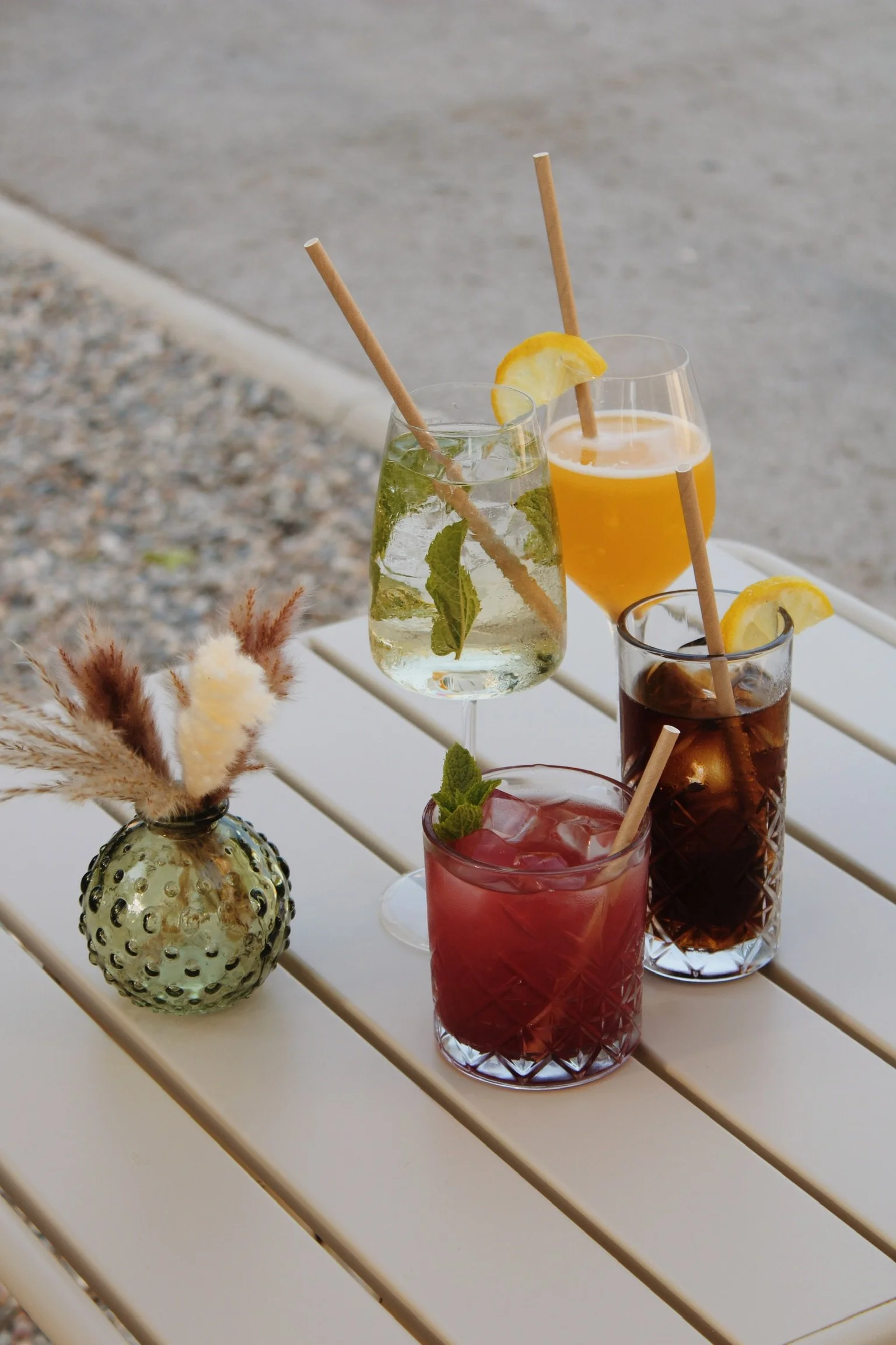 Quatre verres de boissons colorées avec des tranches de citron et des pailles en bois sur une table blanche, avec une petite vase décorative contenant des fleurs séchées en premier plan.