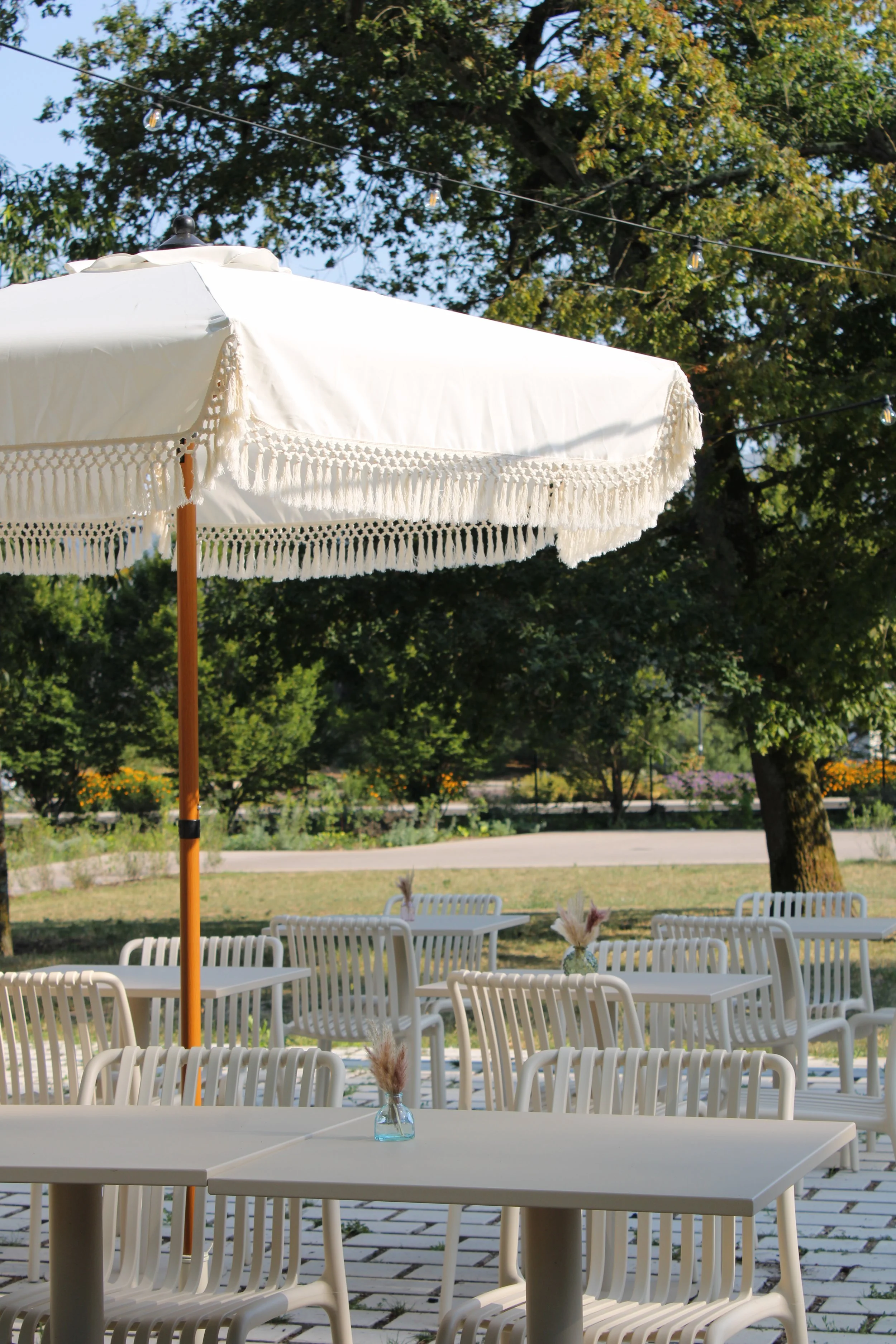 Une grande table blanche en plastique sous un parasol blanc avec des bords en crochets de coton, dans un espace extérieur avec des chaises blanches, des fleurs dans des vases en verre, des arbres et un ciel bleu en arrière-plan.