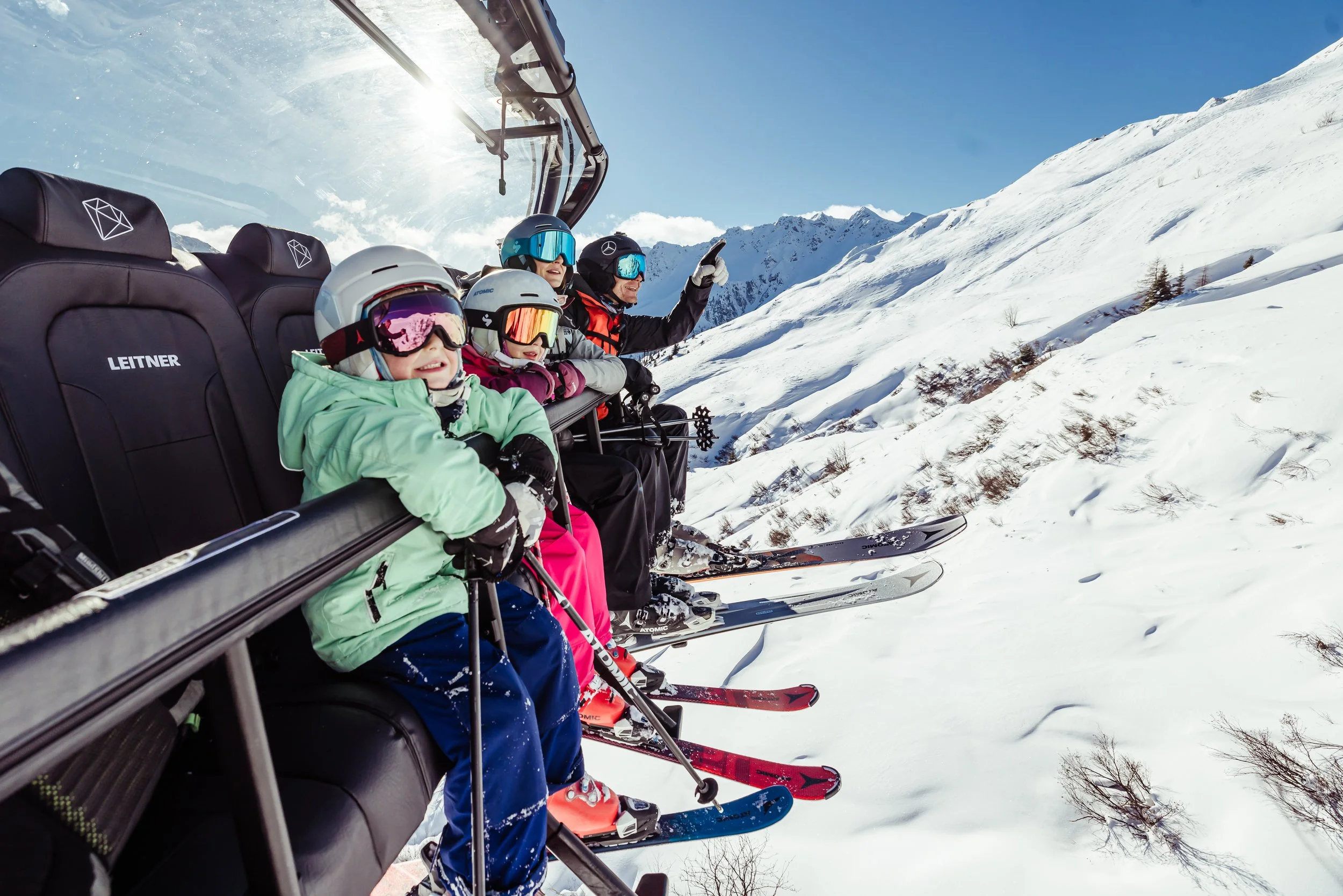Vier Personen, einschließlich Kinder, sitzen in einem Skilift in verschneiten Bergen, tragen Ski- und Schneekleidung sowie Helme und Skibrillen, während die Sonne am klaren Himmel scheint.