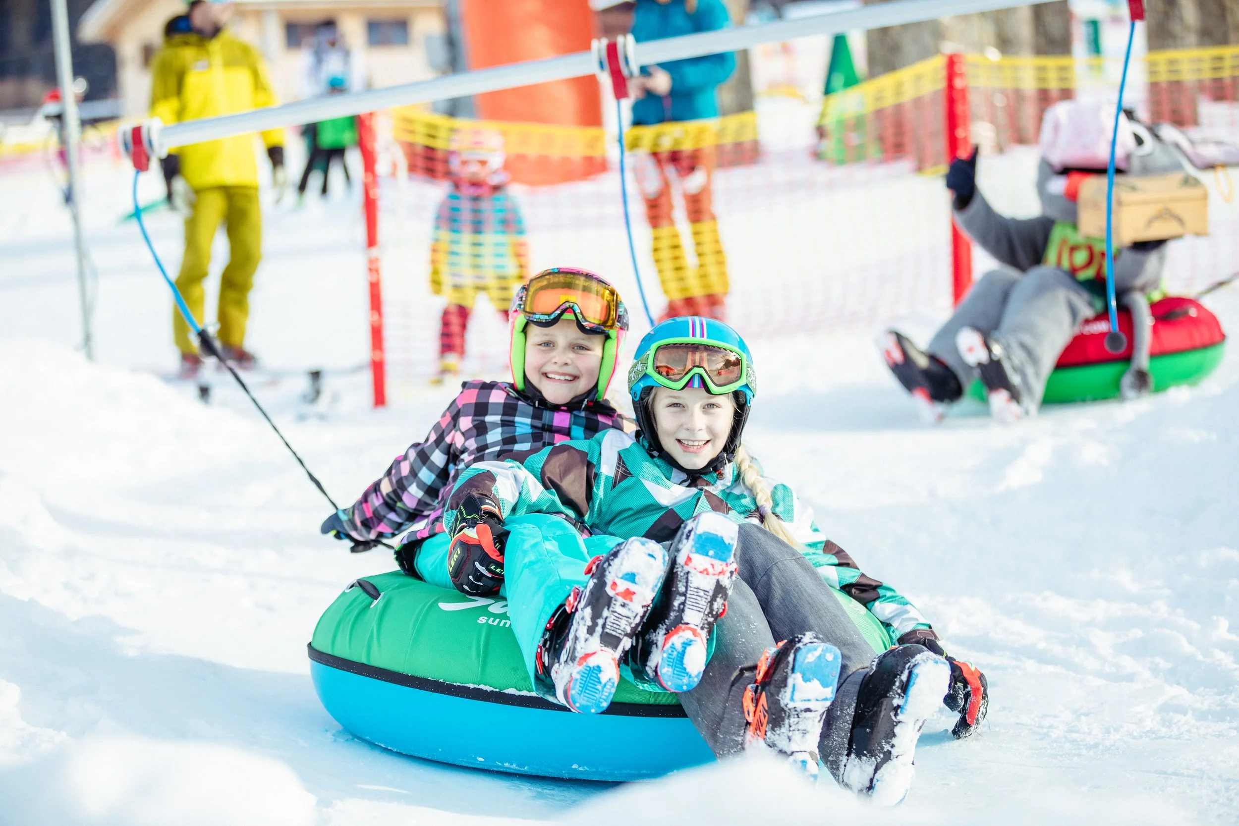 Zwei lachende Kinder beim Rodeln im Schnee, beide tragen Helme und Wintersportausrüstung. Im Hintergrund stehen weitere Kinder und Erwachsene, sowie Sicherheitszäune auf einer Skipiste.