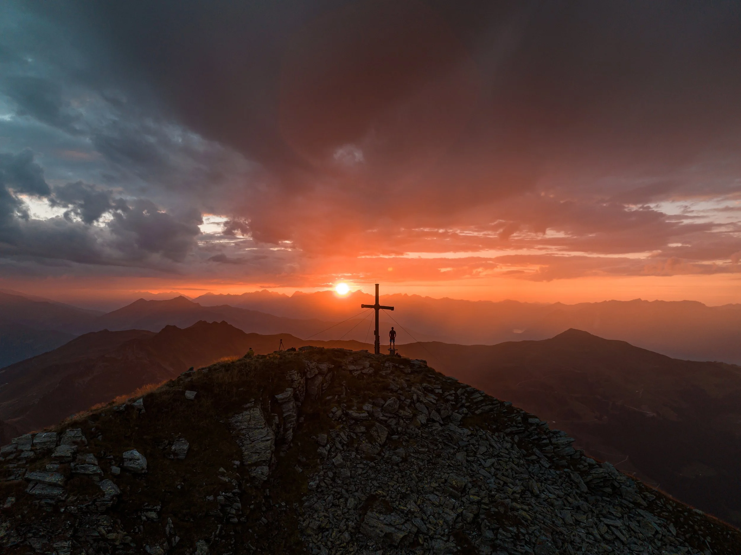 Ein Berggipfel bei Sonnenuntergang mit einem Holzkreuz und einer Person, die neben dem Kreuz steht. Im Hintergrund sind Berge und ein bewölkter Himmel mit dunklen und orangefarbenen Wolken.