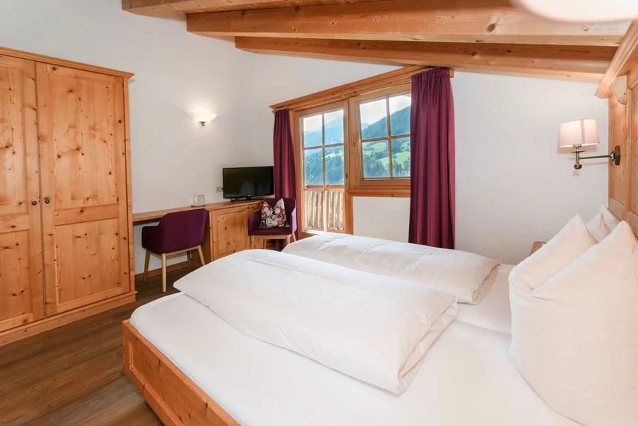 Ein Zimmer mit einem Doppelbett, Holzmöbeln, einer Fensterfront mit Bergblick, einem Fernseher, einem Stuhl, einem Schreibtisch und roten Vorhängen.