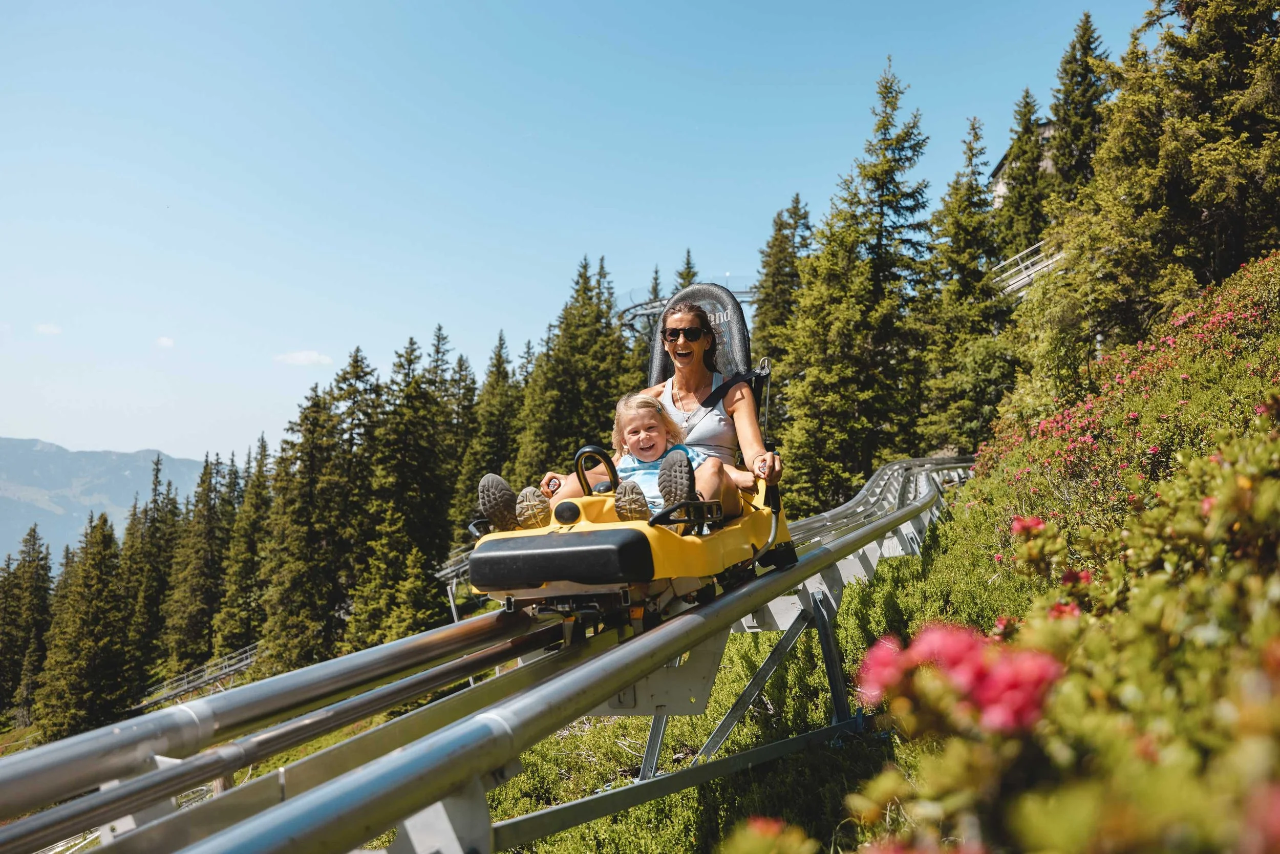 Eine lachende Mutter und ihr Kind fahren in einer Sommerrodelbahn im Grünen bei schönem Wetter