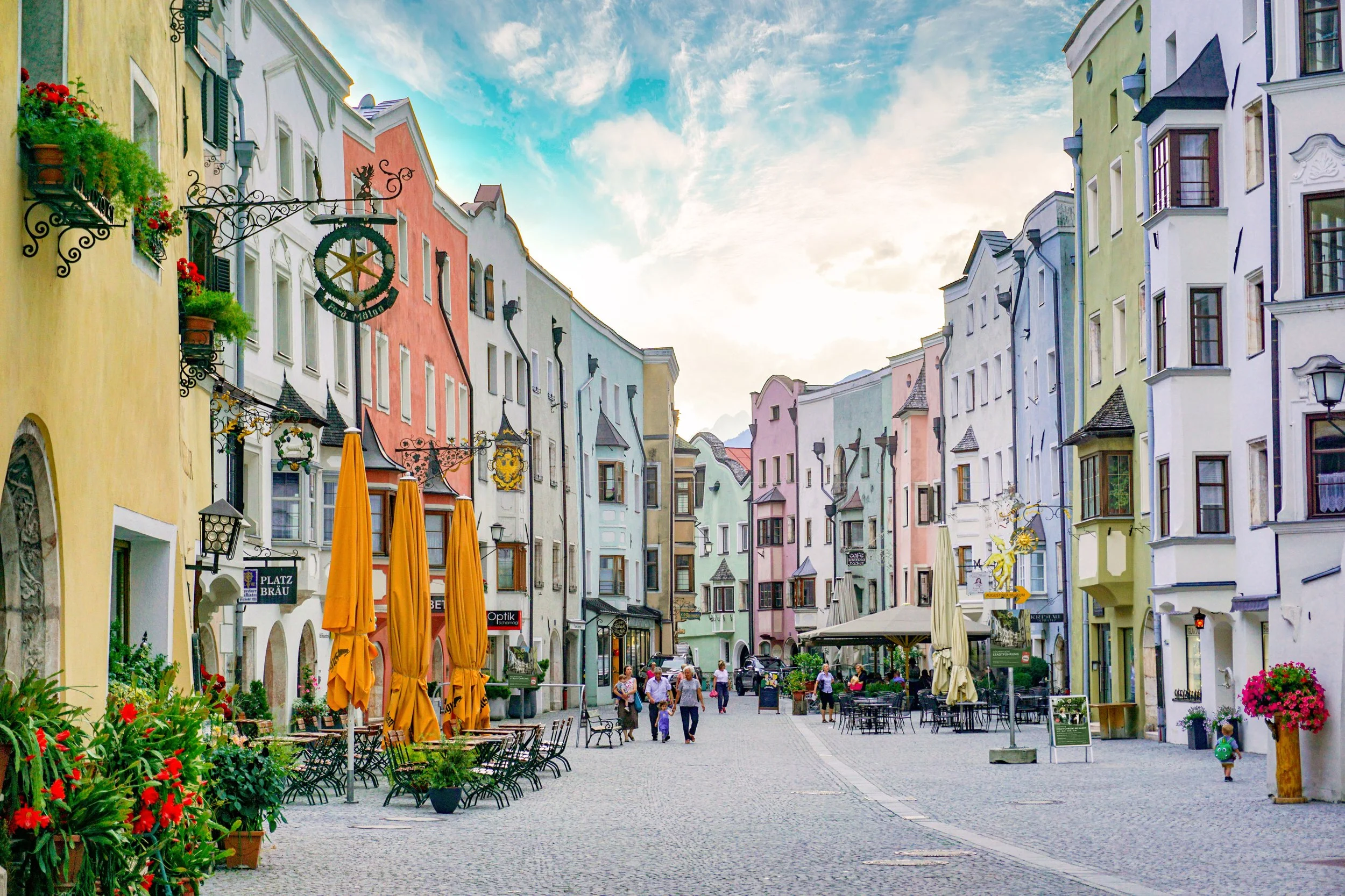 Bunte Häuser in einer Fußgängerzone mit Tischen und Stühle im Freien, Menschen spazieren, einige sitzen an Tischen, blühende Pflanzen, helle Tageszeit.