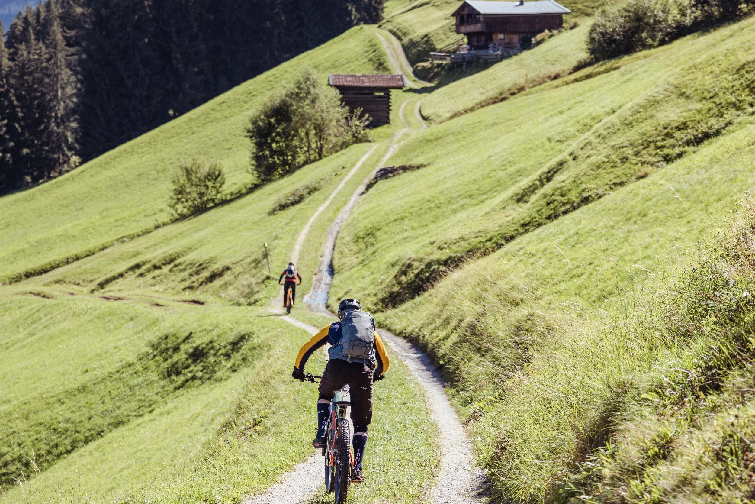 Zwei Mountainbiker auf einem schmalen Wanderweg in einer grünen, bergigen Landschaft mit Hütten im Hintergrund.