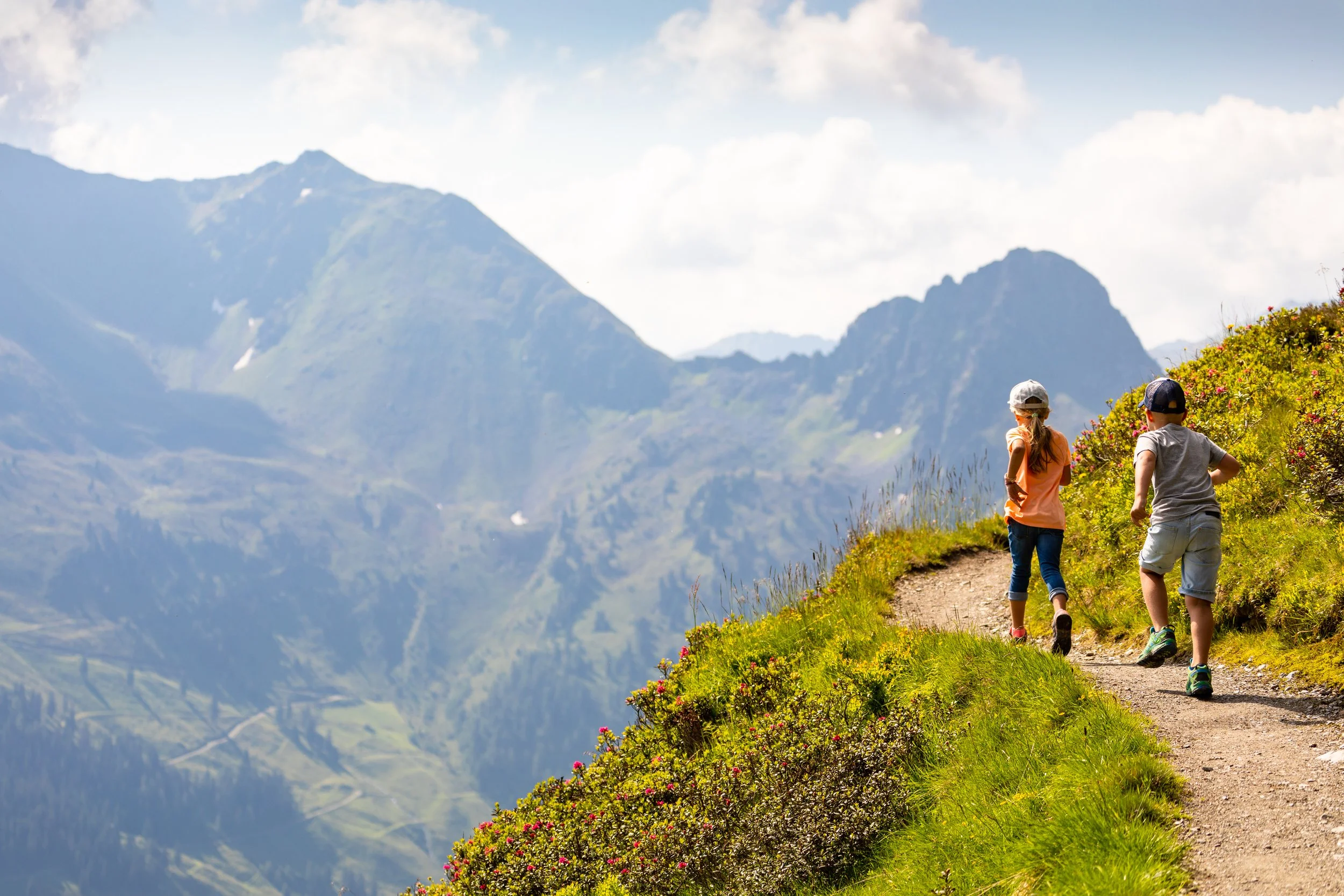 Zwei Kinder wandern auf einem schmalen Weg in einer bergigen, grünen Landschaft mit Bergen im Hintergrund.