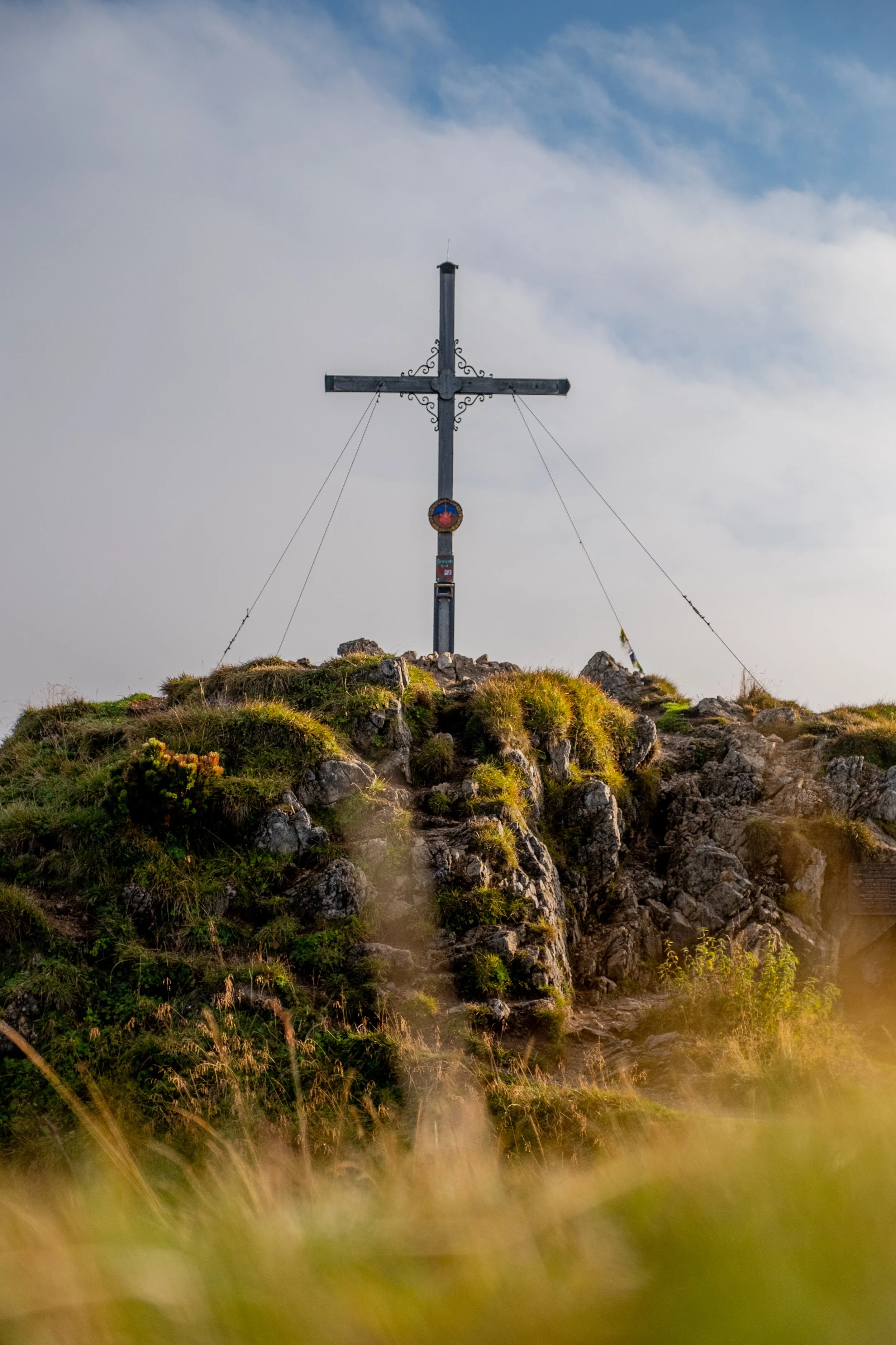 Metallkreuz auf einem felsigen Hügel mit Gras im Vordergrund, Himmel mit Wolken im Hintergrund.