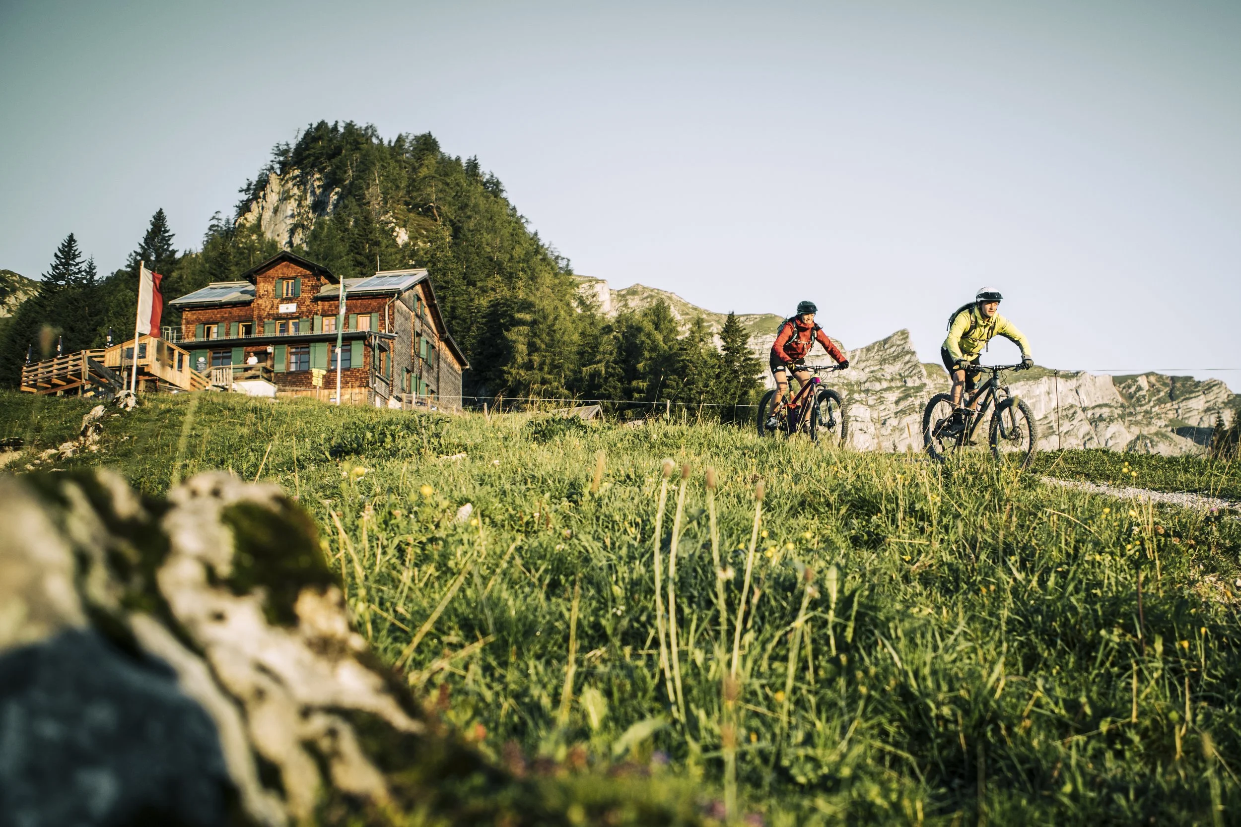 Zwei Mountainbiker fahren auf einer grünen Wiese in den Bergen. Im Hintergrund sieht man ein Holzhaus mit Solarzellen auf dem Dach, umgeben von Bäumen und Felsen, bei Tageslicht.