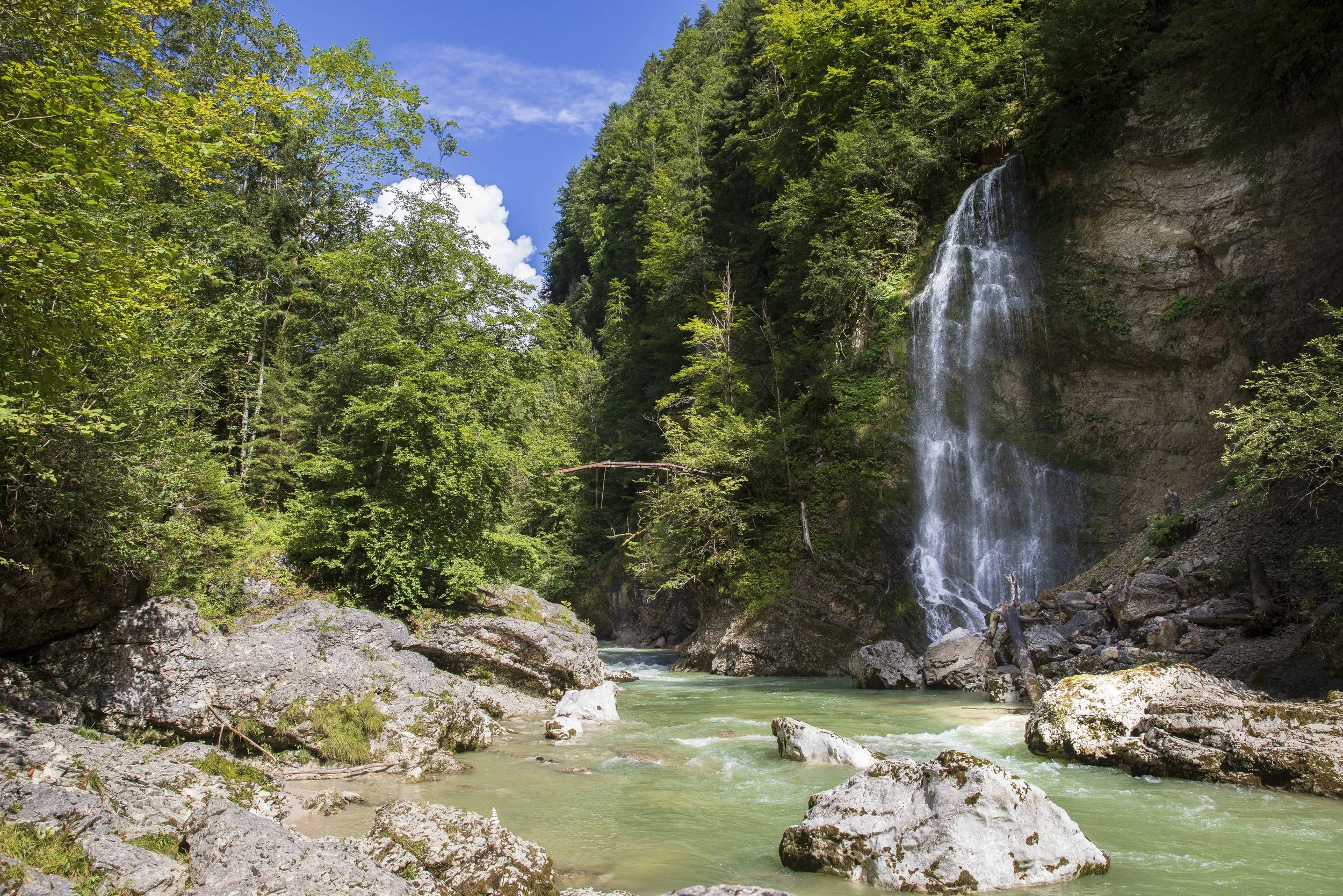 Ein Wasserfall fließt an einer felsigen Klippe in einen Fluss, umgeben von grünen Bäumen unter einem blauen Himmel.
