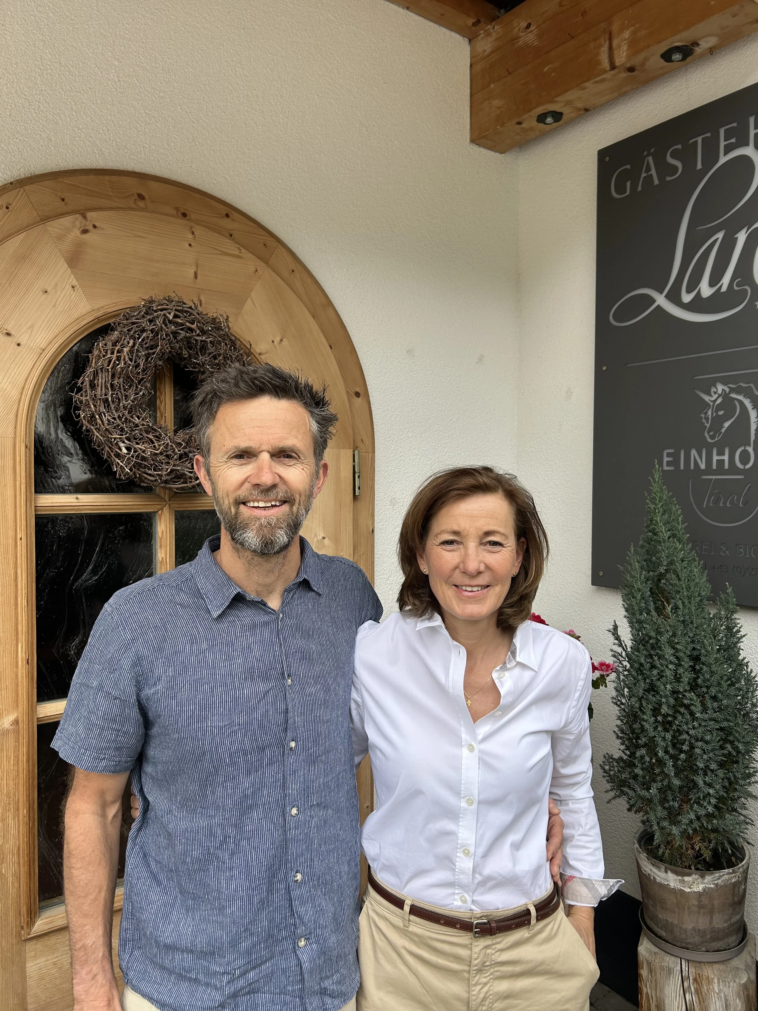 Ein Mann und eine Frau stehen nebeneinander vor einer Tür mit einer Kranzgirlande, daneben ein Bonsai. Es scheint sich um ein Restaurant oder Gasthaus zu handeln.